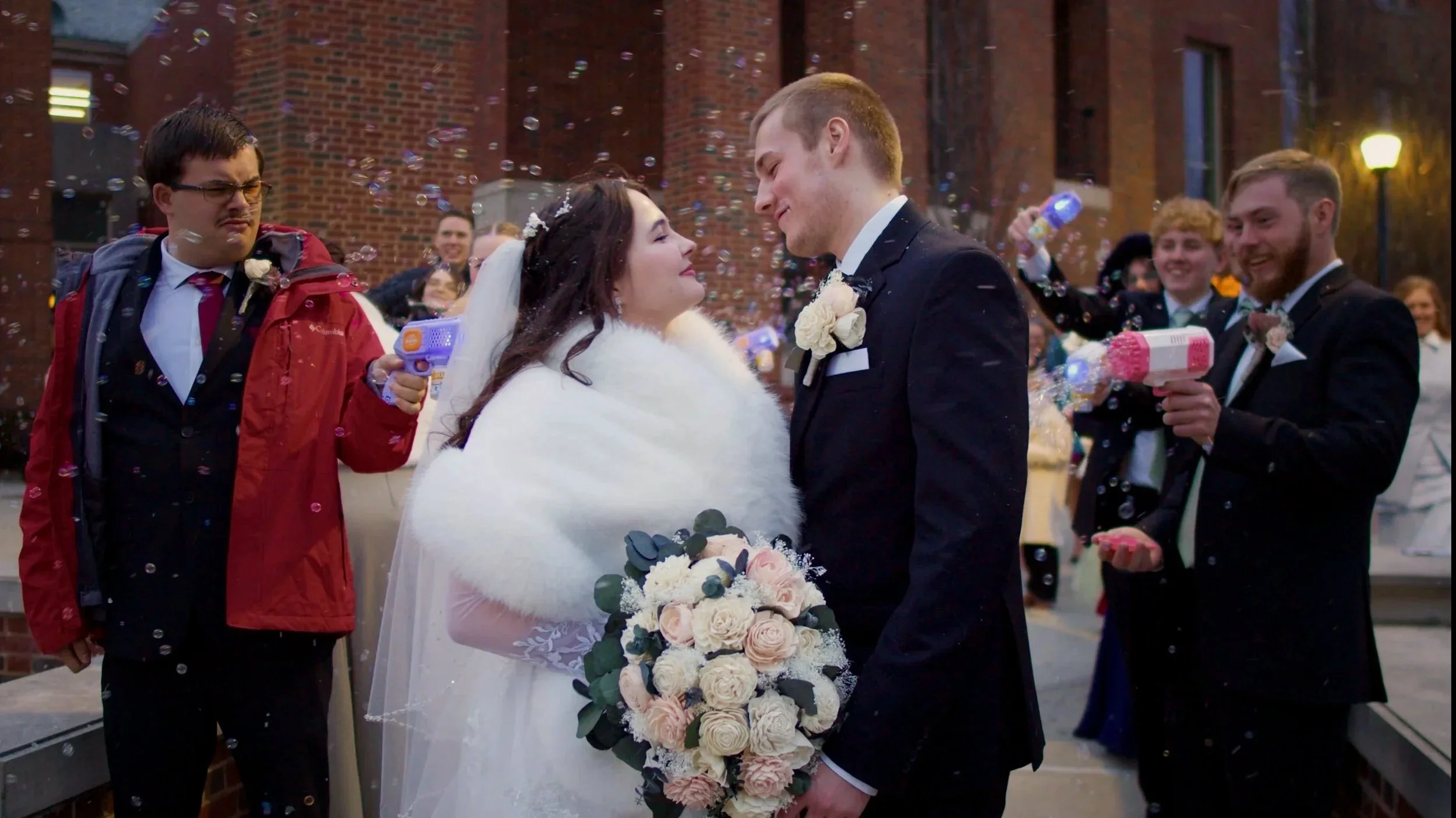 A bride and groom in wedding attire staring at each other surrounded by friends and family celebrating with bubbles outside.