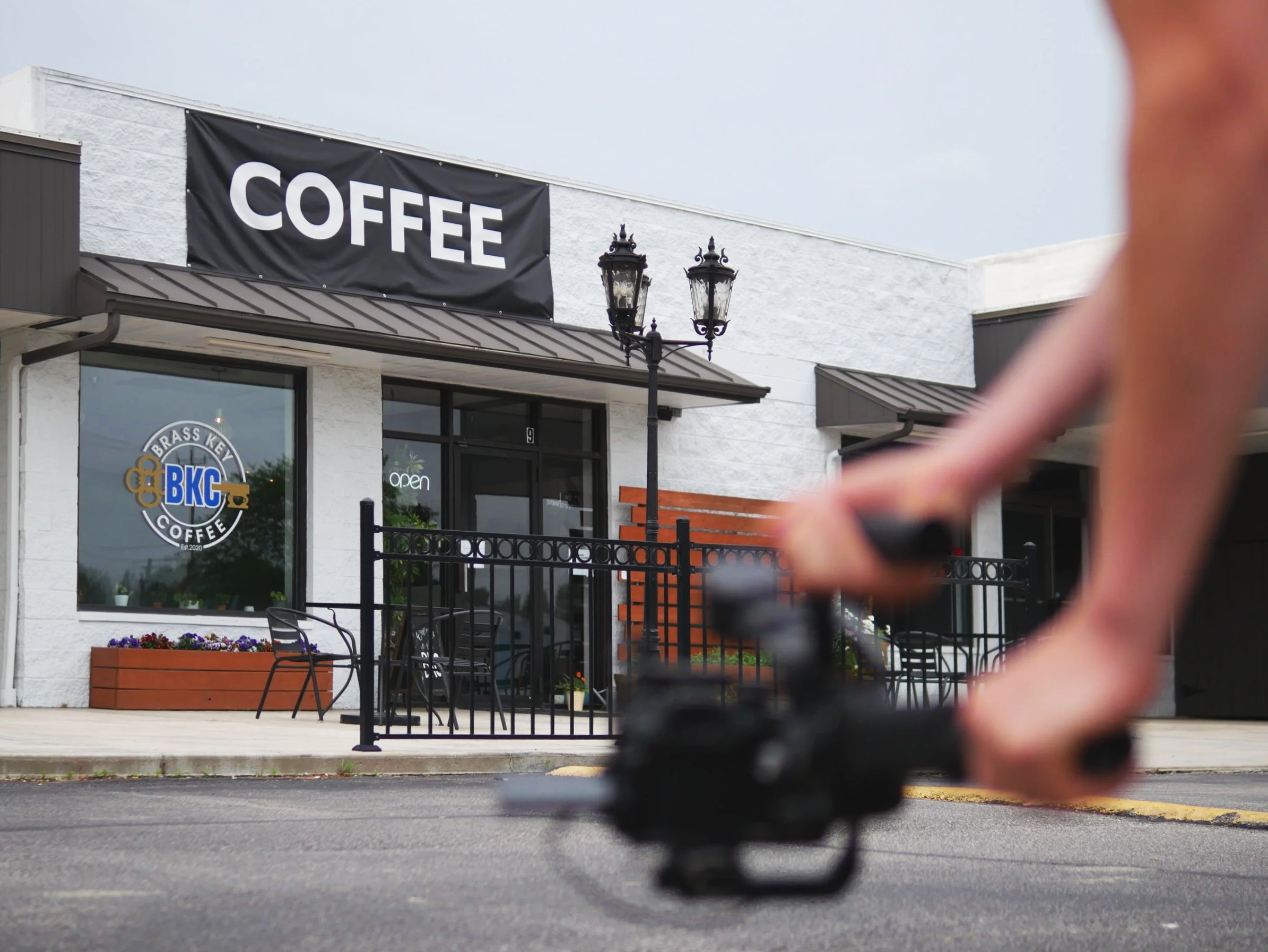 Close-up of a person holding a camera, capturing video of the outside of a coffee shop called Brass Key Coffee with sign that says 'COFFEE', with outdoor seating and a street lamp in front of the shop.
