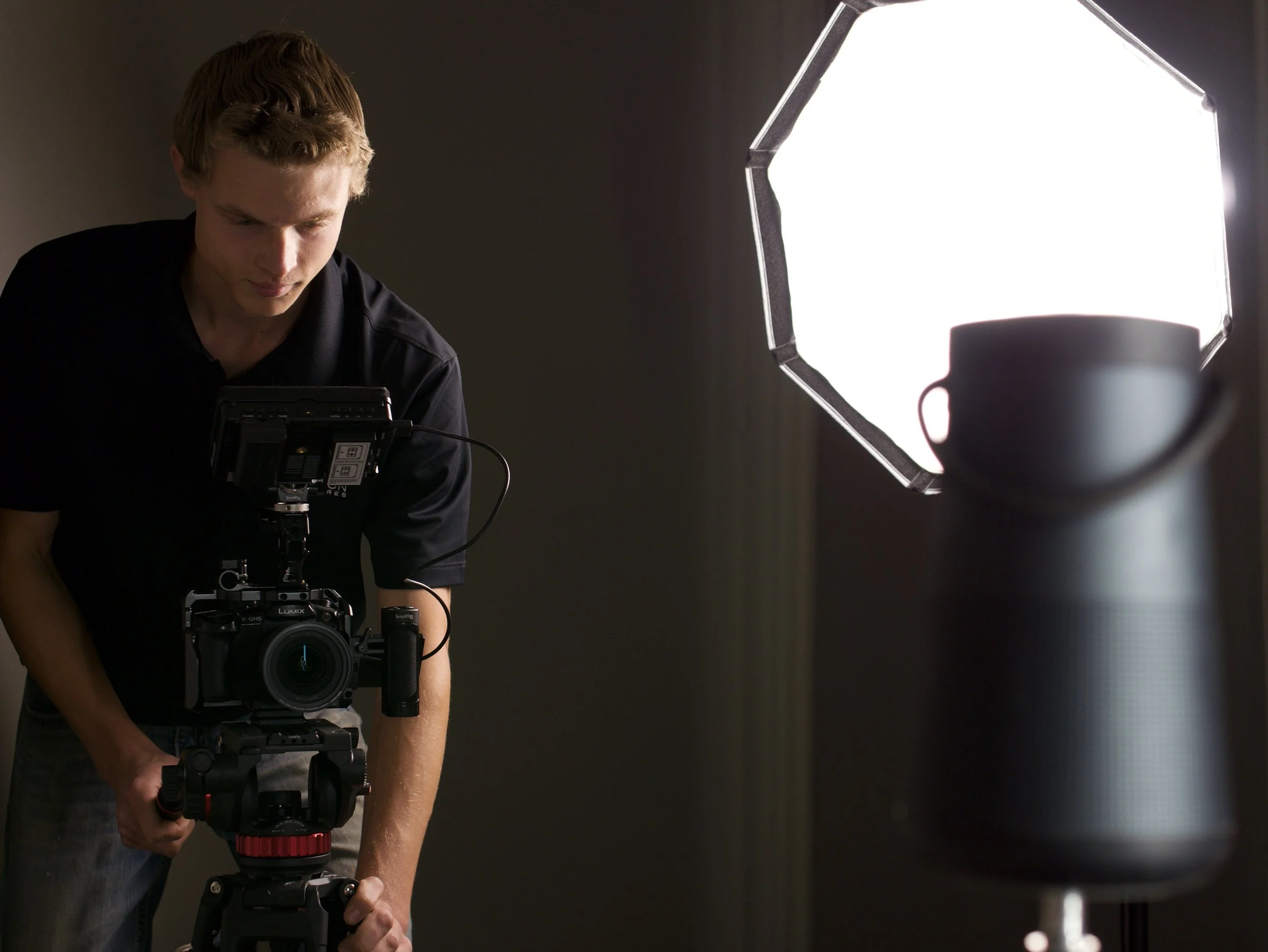 A man operating a professional camera on a tripod in a dark room, with a large bright light source to the right.