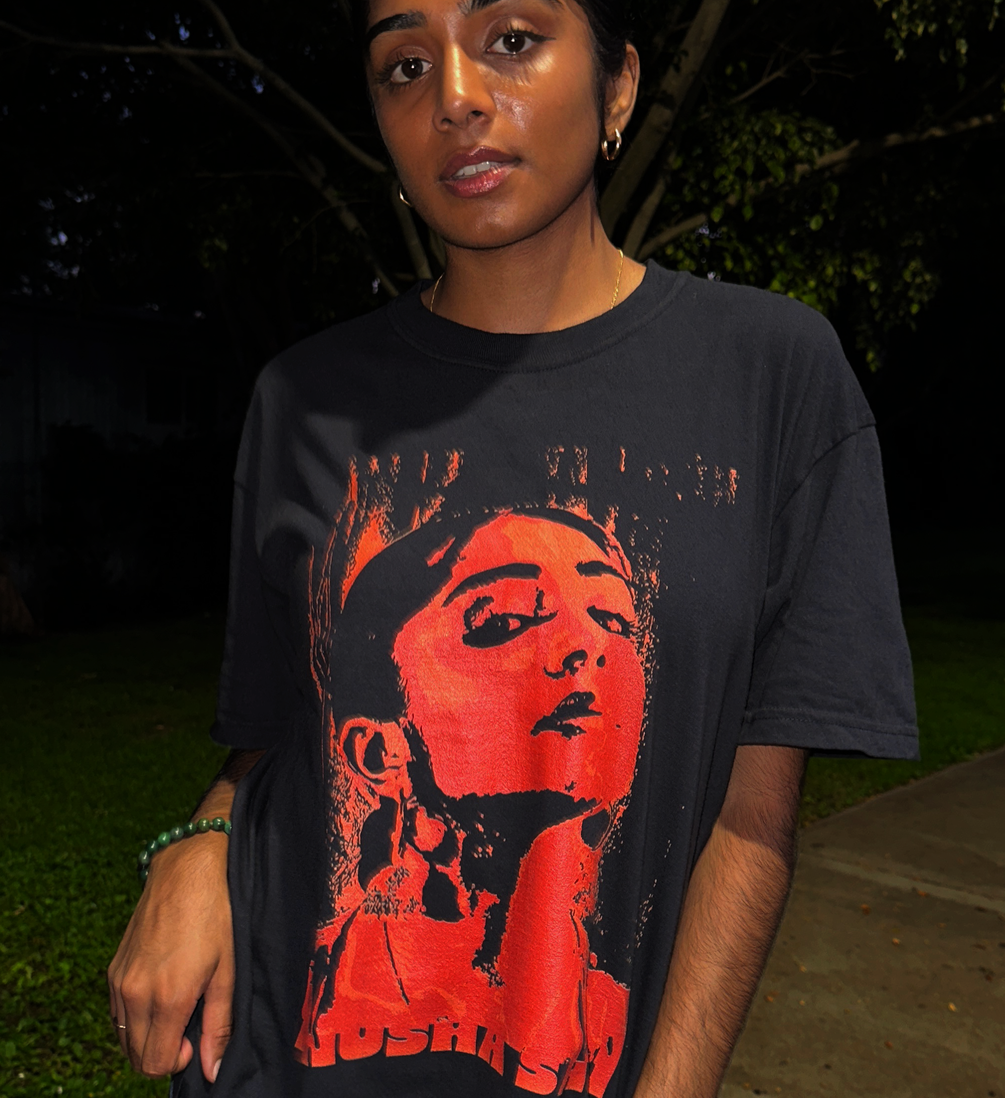 A woman with dark hair and earrings stands outdoors at night, wearing a black T-shirt featuring a graphic of a woman with a bun hairstyle in red and orange tones and the word 'Hustle' at the bottom. She has a beaded bracelet on her wrist and is looki