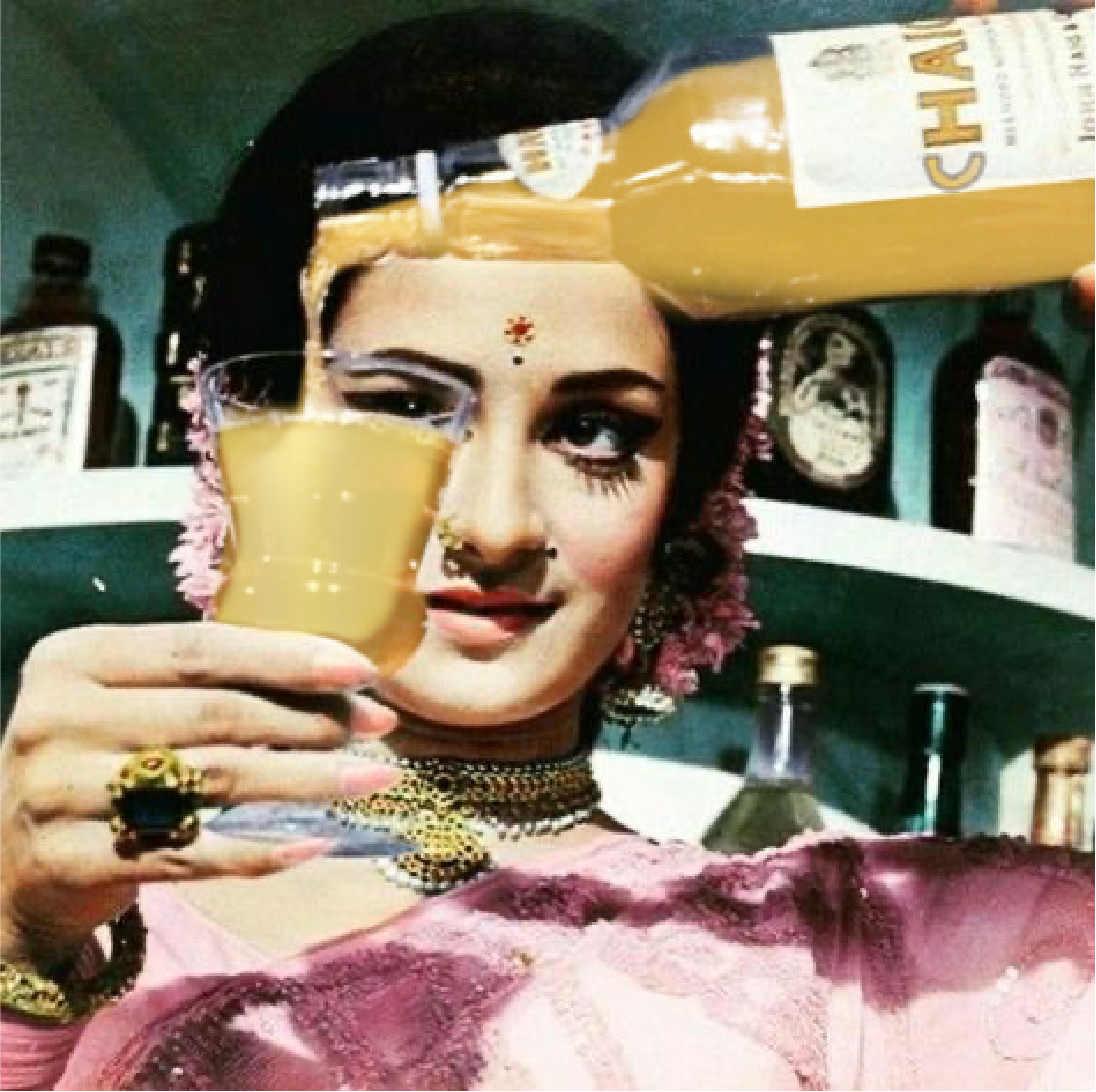 A woman dressed in traditional Indian attire holding a glass of yellow-colored beverage, with jewelry and flowers in her hair, in an indoor setting with bottles and posters in the background.