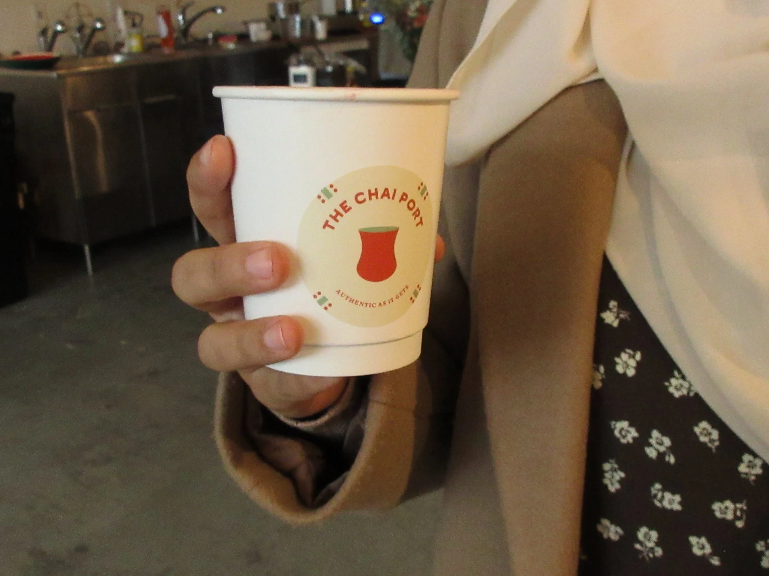 A person holding a white paper cup with a logo that reads 'The Chai Port' and a picture of a chai cup, with a kitchen background.