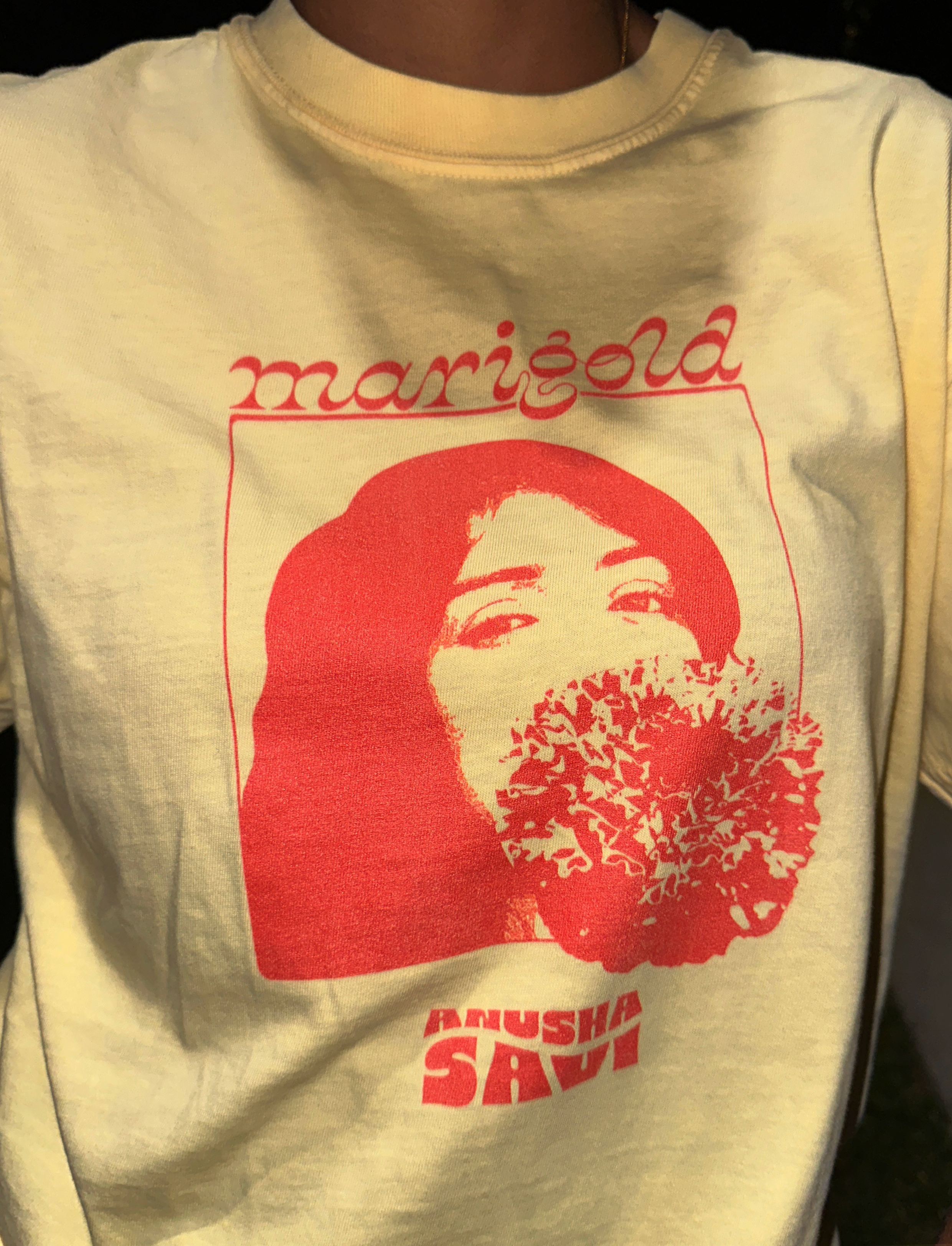 A person wearing a yellow T-shirt with a red graphic of a woman's face that is made up of a flower, with the word 'Marie' written above and partially visible red text at the bottom. The person is also wearing a gold chain and has a dark background.