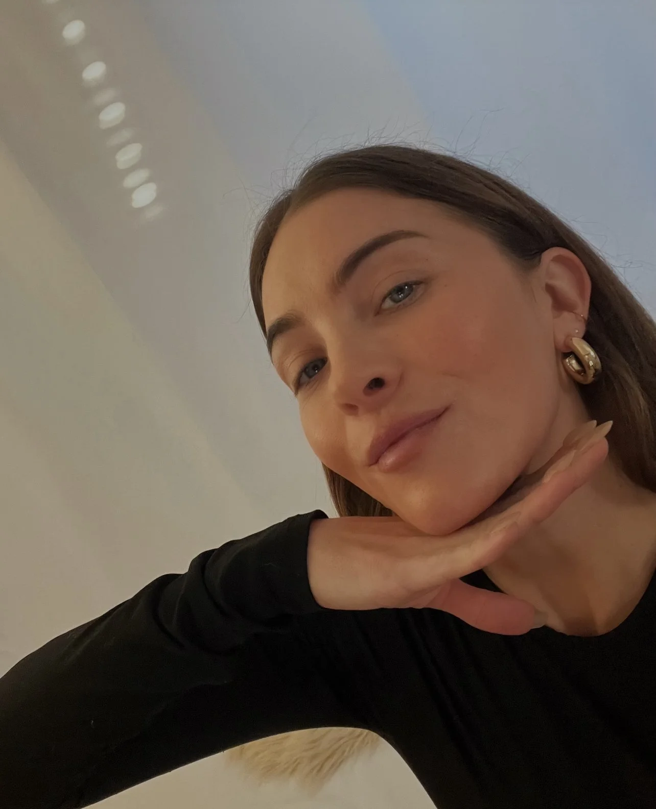 Close-up of a young woman with brown hair, wearing gold earrings, resting her chin on her hand, in an indoor setting.