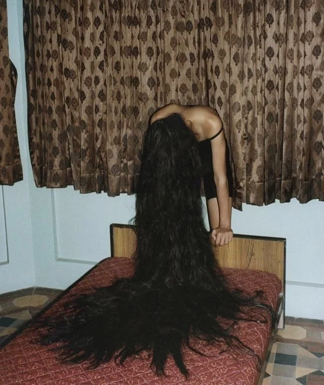 Person bending forward on a bed with extremely long black hair cascading across the bedspread