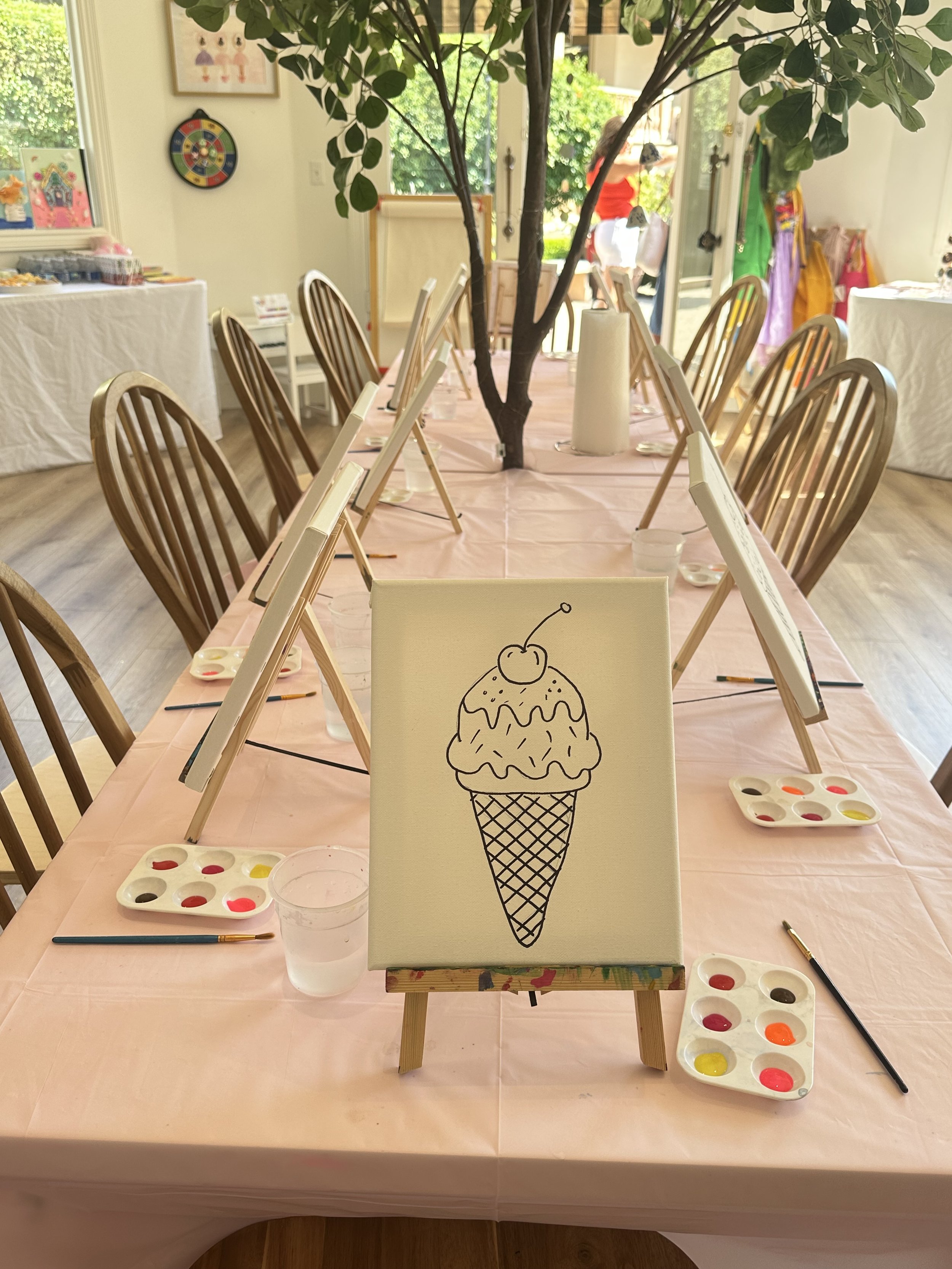 A table for a painting activity with paint palettes, brushes, and a canvas featuring a line drawing of an ice cream cone with a cherry on top.