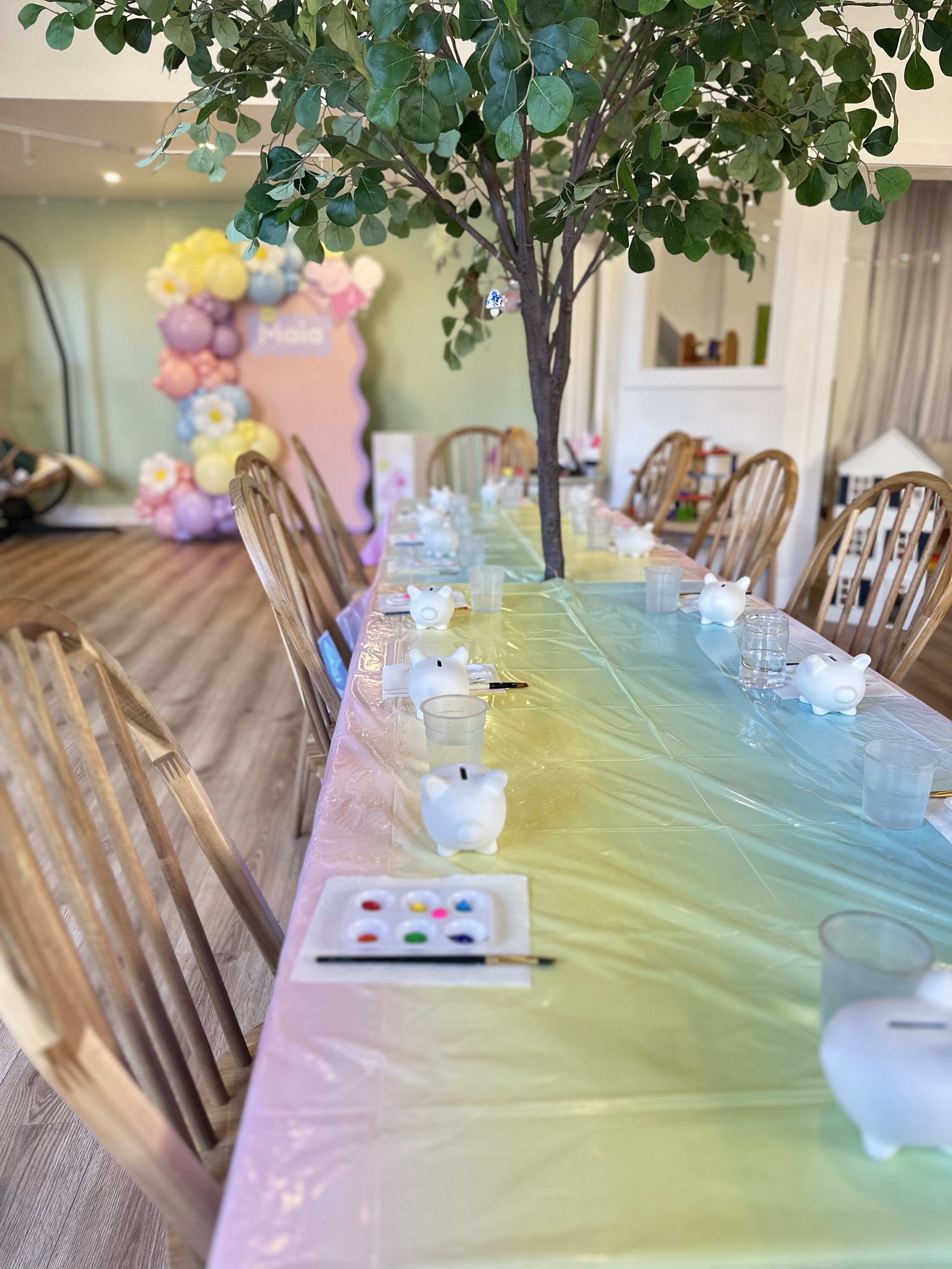 Table decorated for a celebration with a large artificial tree in the center, surrounded by yellow, pink, and purple balloon clusters, and a pink sign with white letters. The table has a yellow tablecloth, white piggy bank containers, water cups, and art supplies.