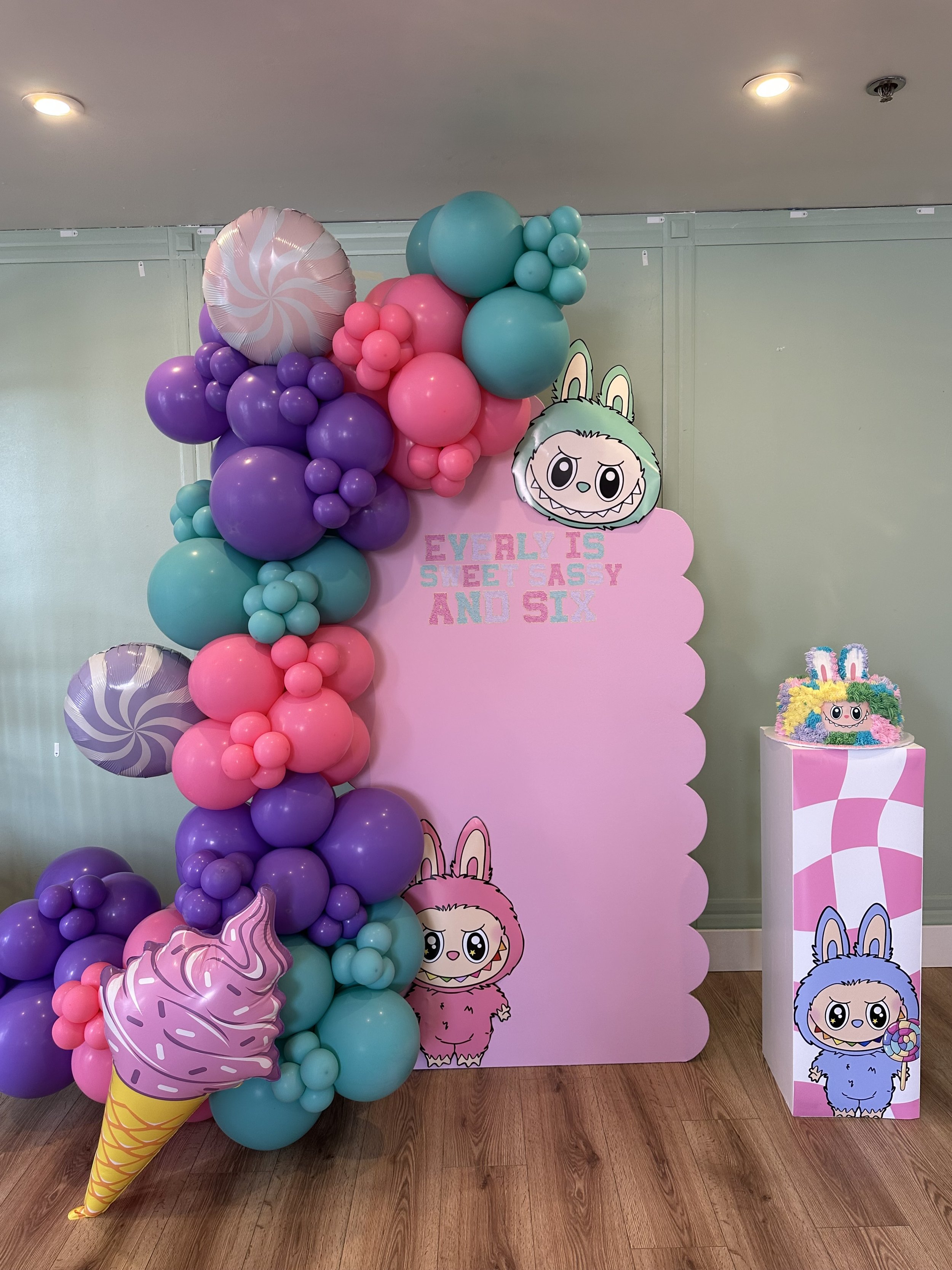 Decorative arrangement with colorful balloons in shades of purple, pink, and teal, including a large ice cream cone balloon and a character cutout with pastel-colored hair and bunny ears, set against a pink backdrop with the words "Every 1 is Sweet Asy and Six" and cartoon characters.
