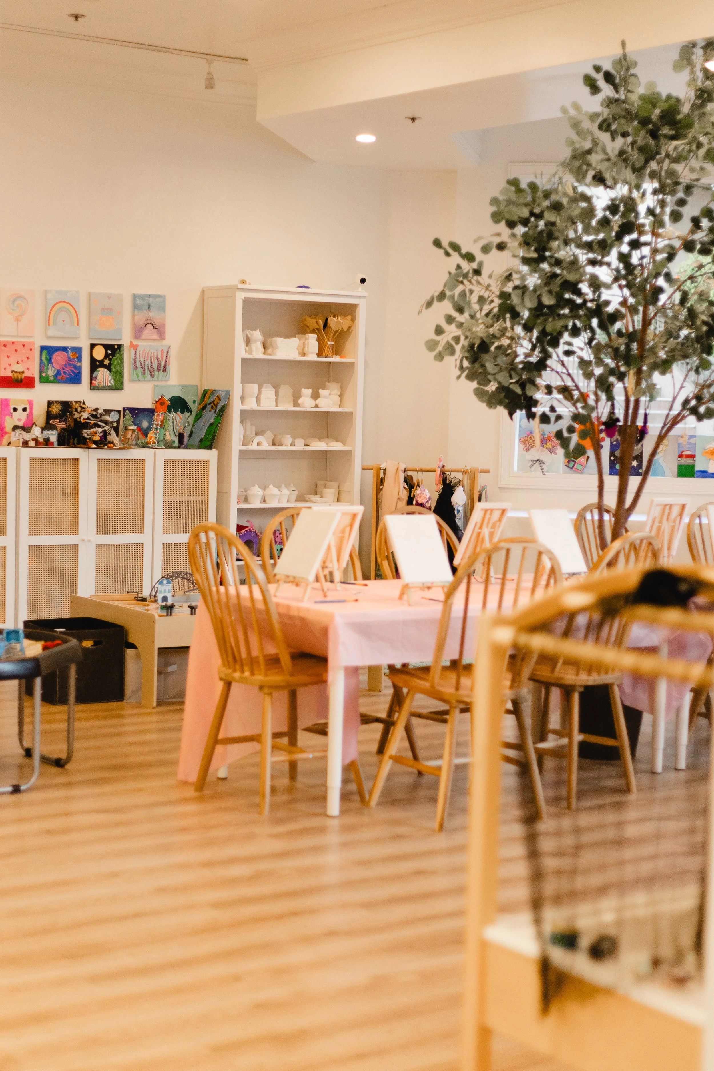 A room decorated with colorful artwork on the wall, a white shelving unit with ceramic figures, and tables with pink tablecloths and wooden chairs. There is a large leafy tree and sunlight coming through the window.