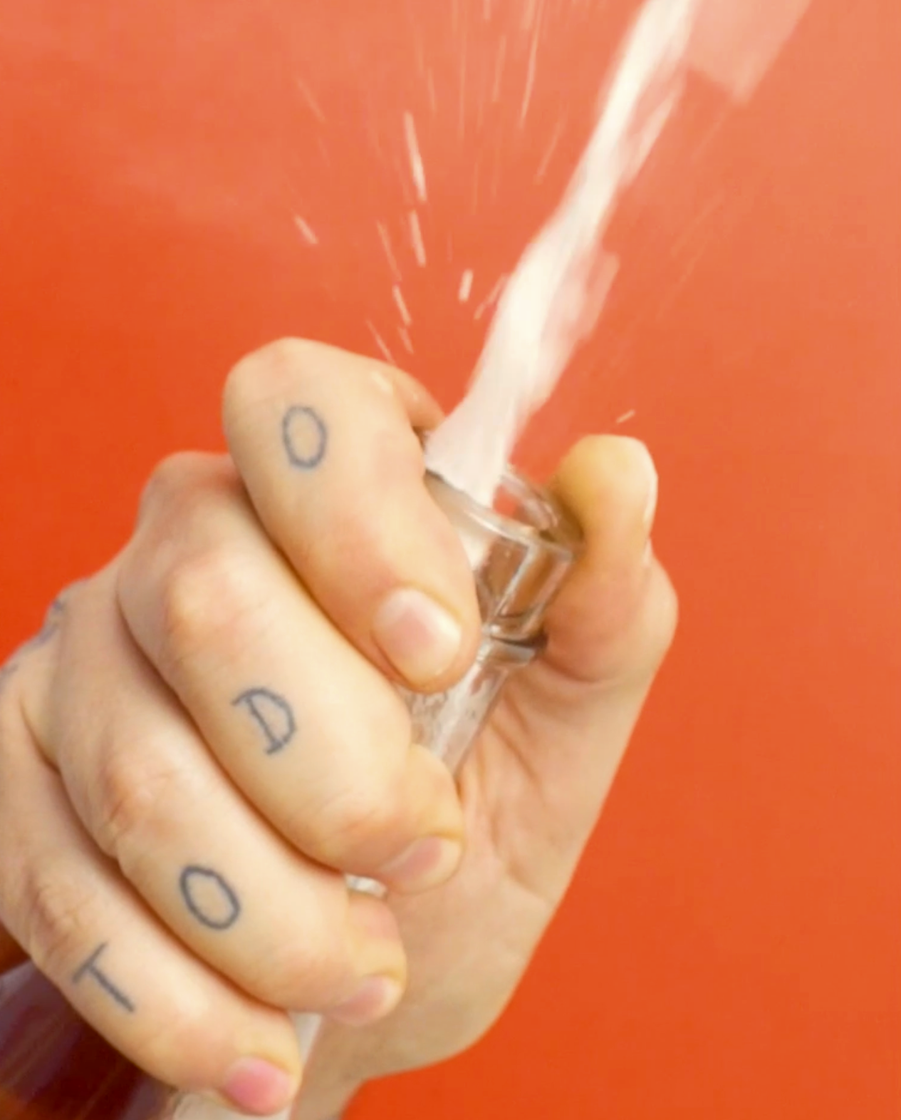 Una mano con tatuajes en los dedos, sosteniendo un aparato de cristal que dispersa polvo o líquido en el aire, con fondo naranja.