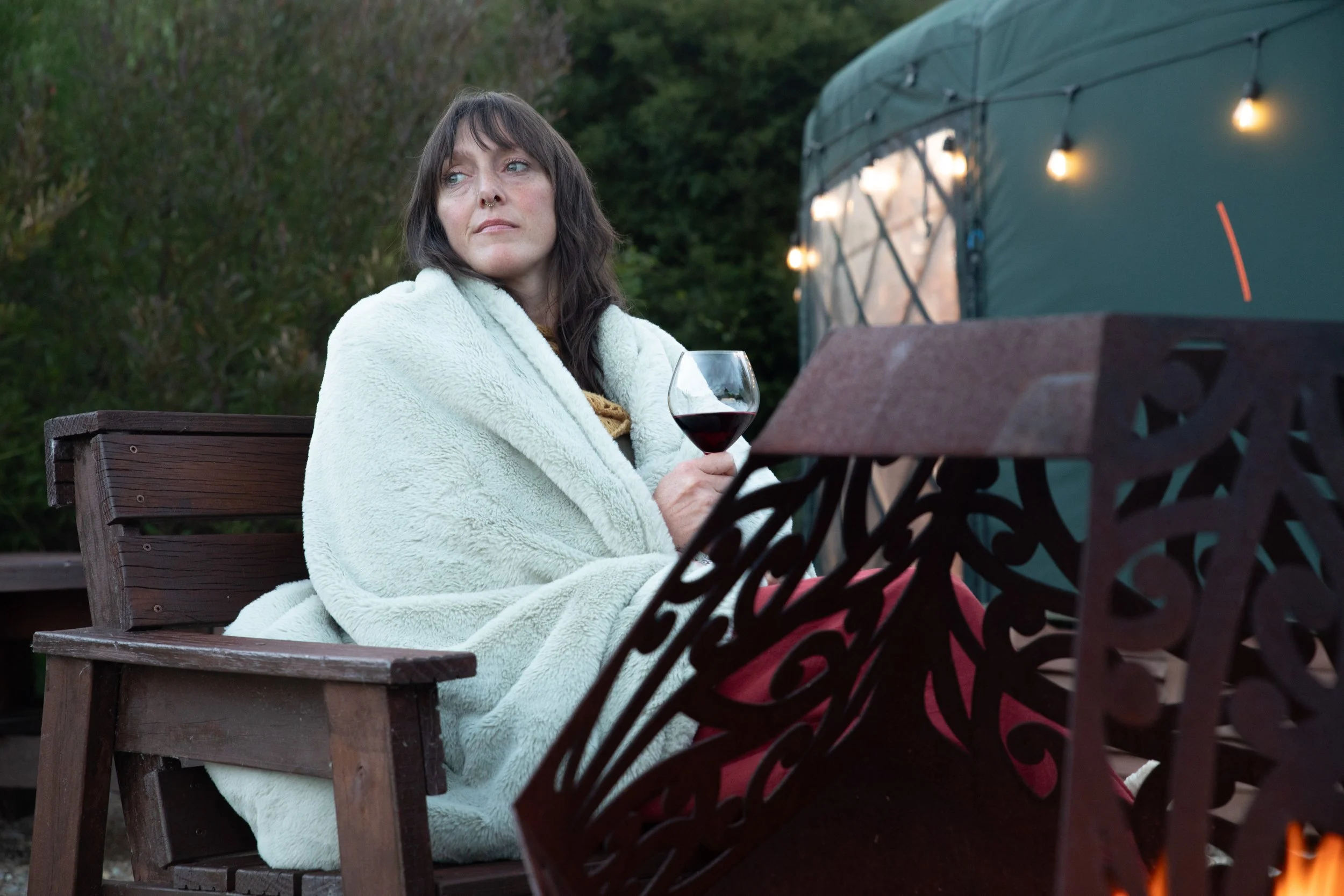 A woman with long dark hair, wearing a cosy, teal blanket, sitting on a wooden garden chair at dusk, holding a glass of red wine, with a yurt and string lights in the background.