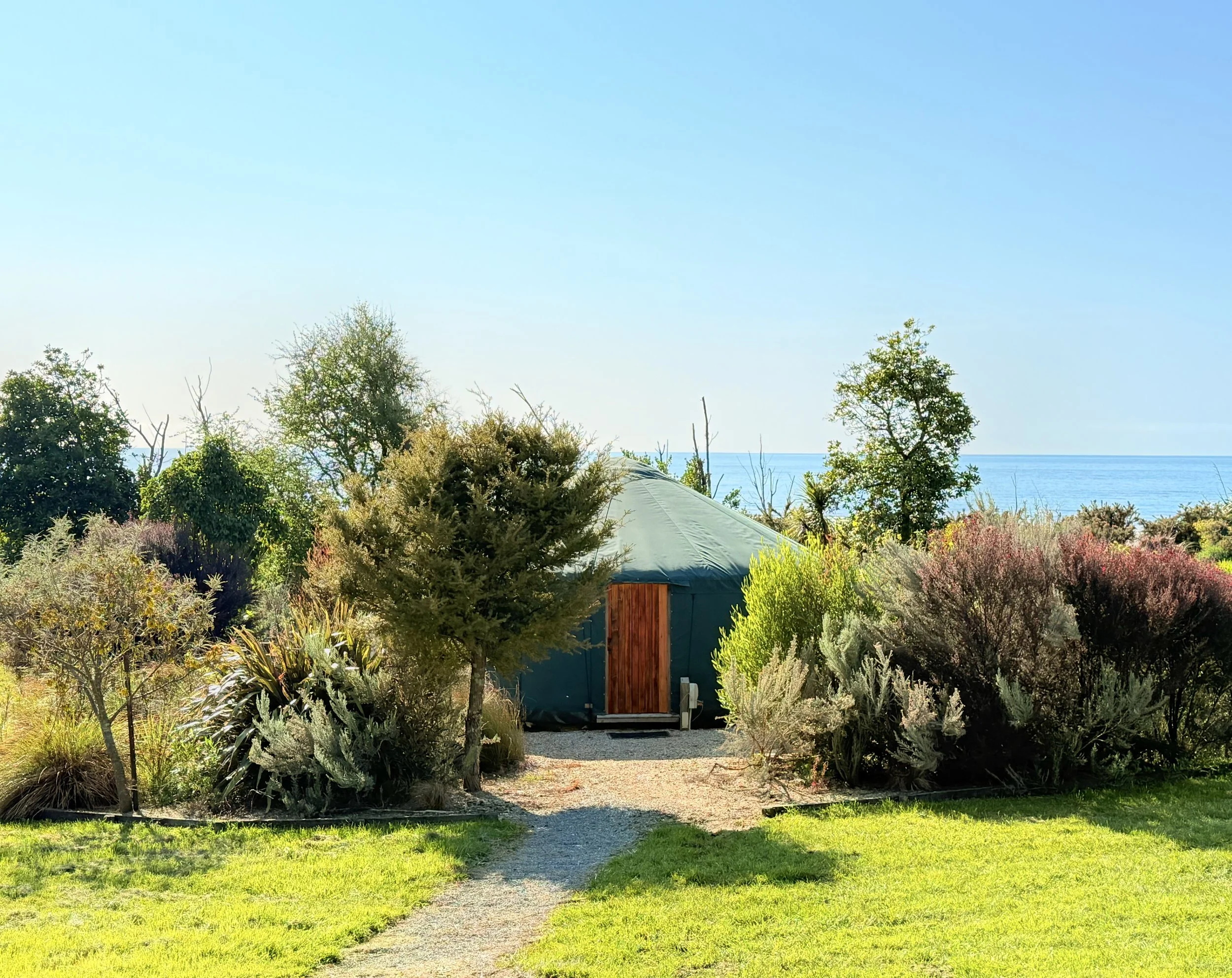 A backyard view with lush green grass, a gravel pathway, various shrubs and trees, a small shed with a rounded roof, and a body of water in the background under a clear blue sky.