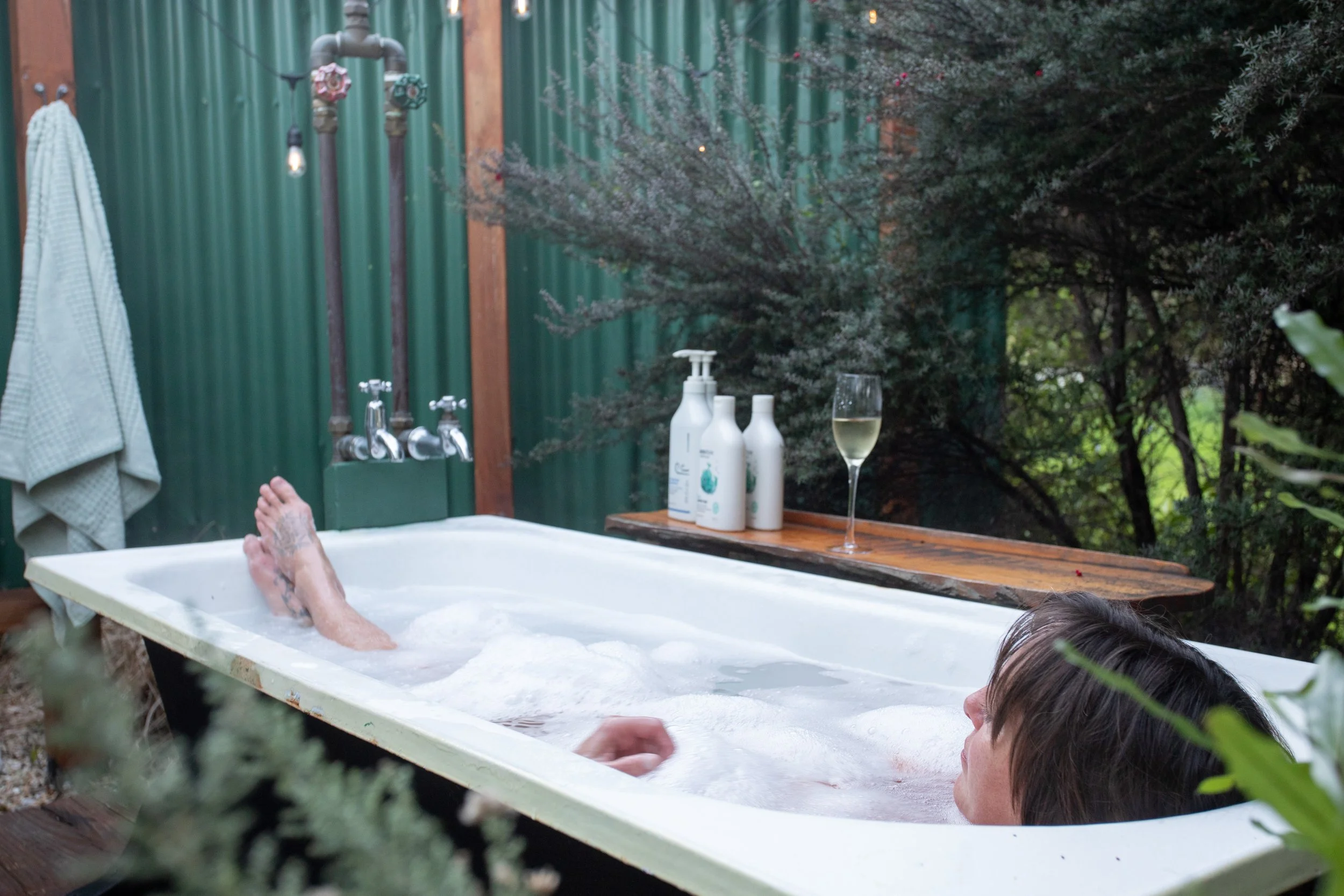 Person relaxing in an outdoor bathtub filled with bubbles, surrounded by greenery, with a glass of sparkling wine on a wooden table nearby.