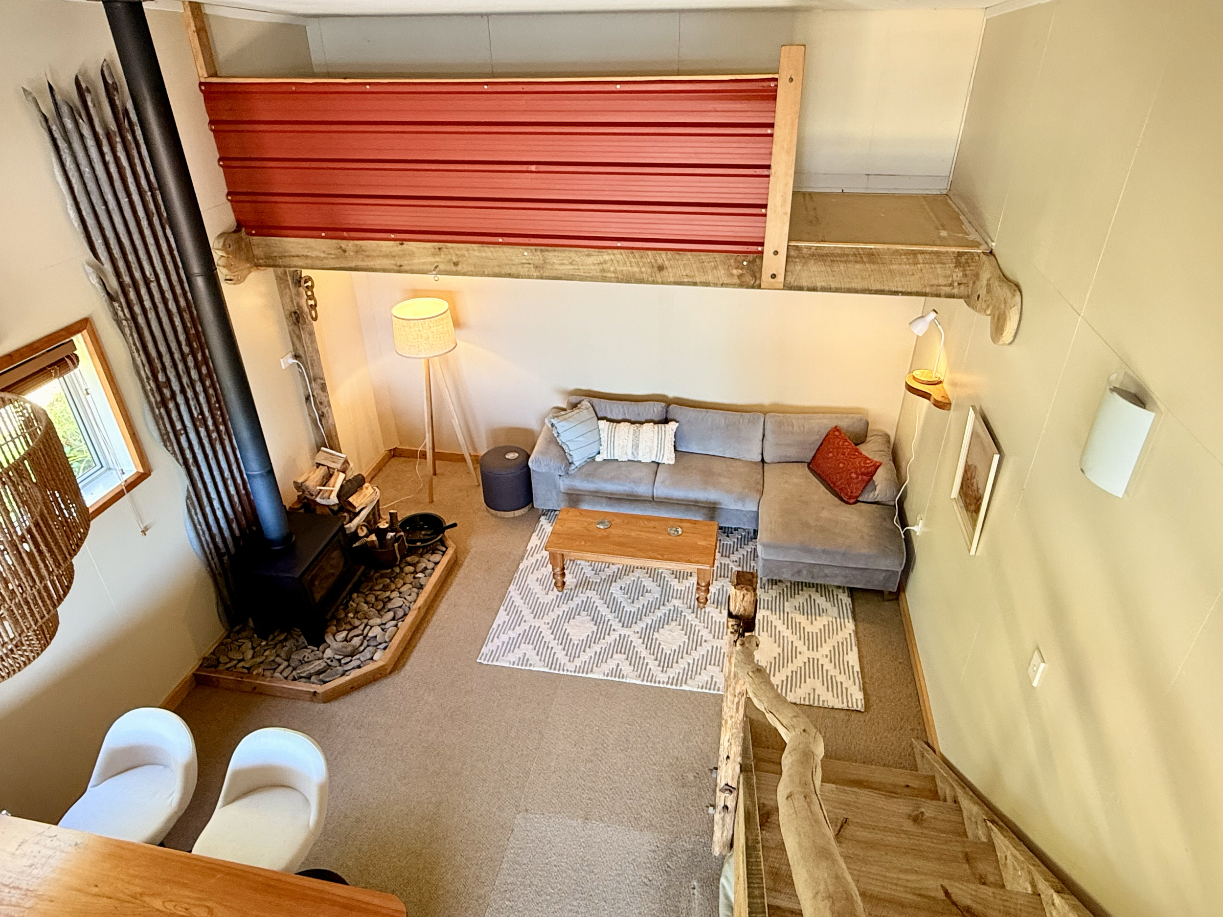Living room with a grey sectional sofa, a wooden coffee table, a patterned rug, a floor lamp, a wood stove with rocks, and a loft with a red railing. There are two white bar stools, a small window, and wall-mounted lamps.