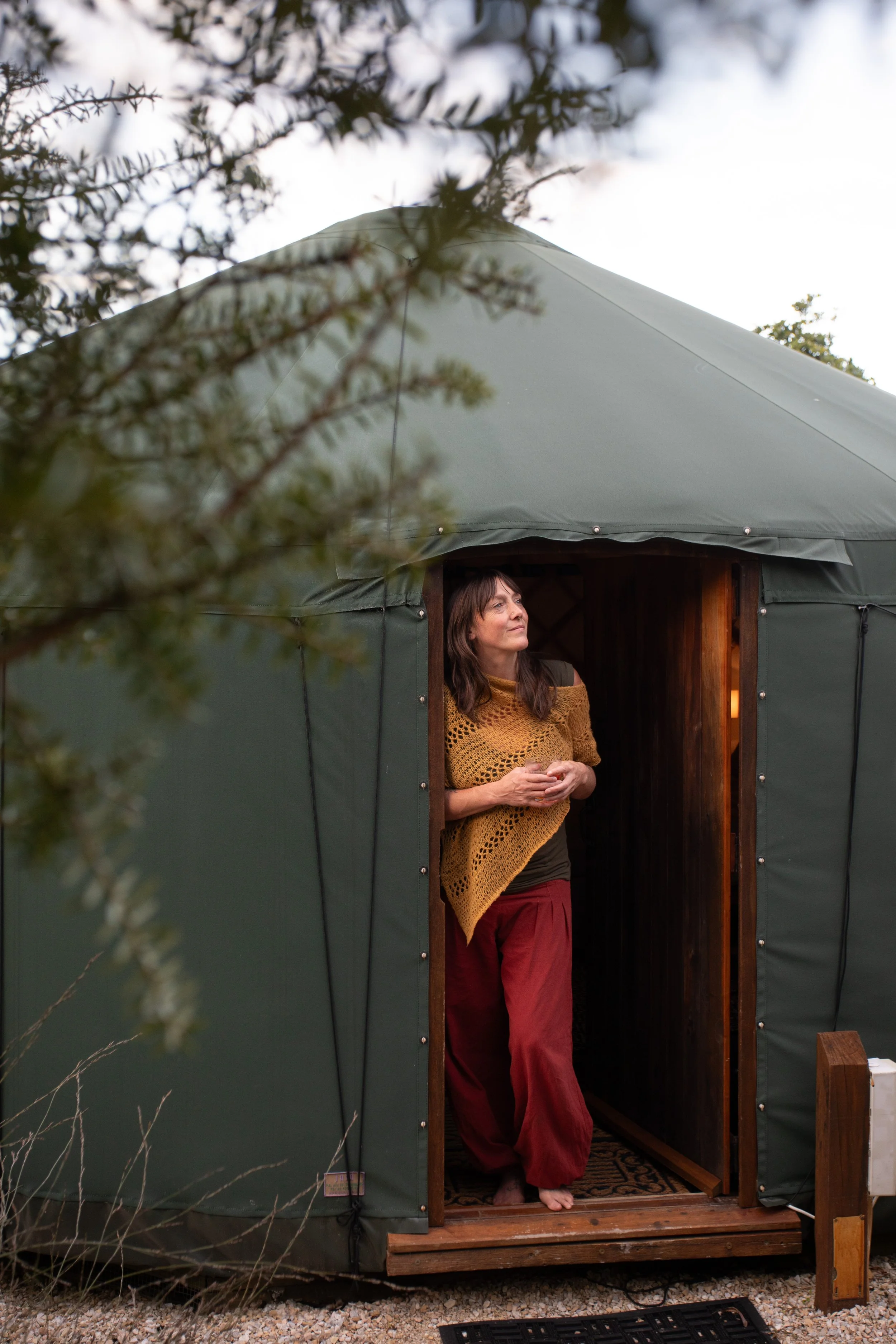 A woman stands barefoot in the doorway of a green, fabric yurt, surrounded by trees and outdoor scenery, with her hands held together.