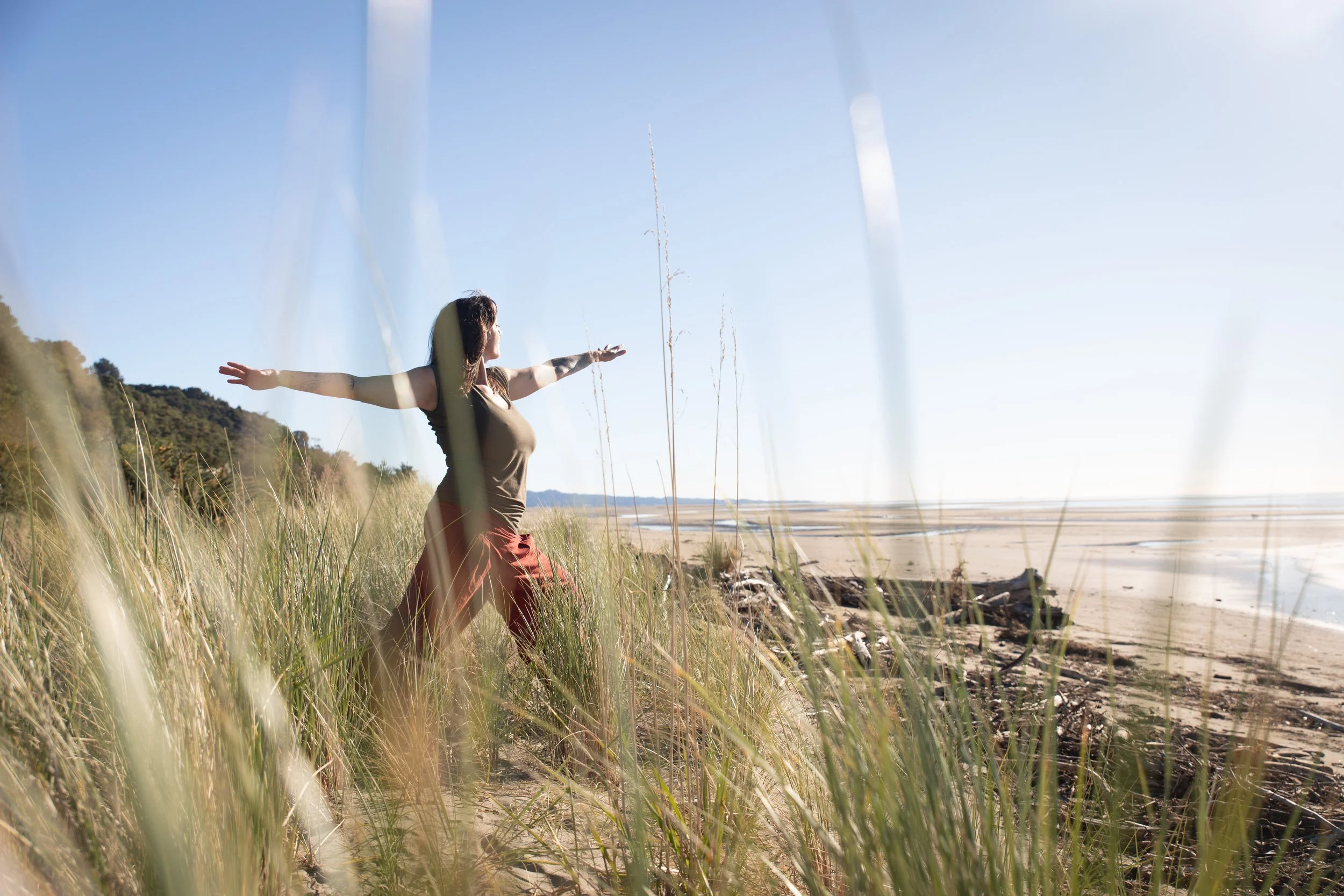 A woman with arms outstretched doing yoga standing in grassy dunes by the beach during daytime, with a clear blue sky and distant hills.