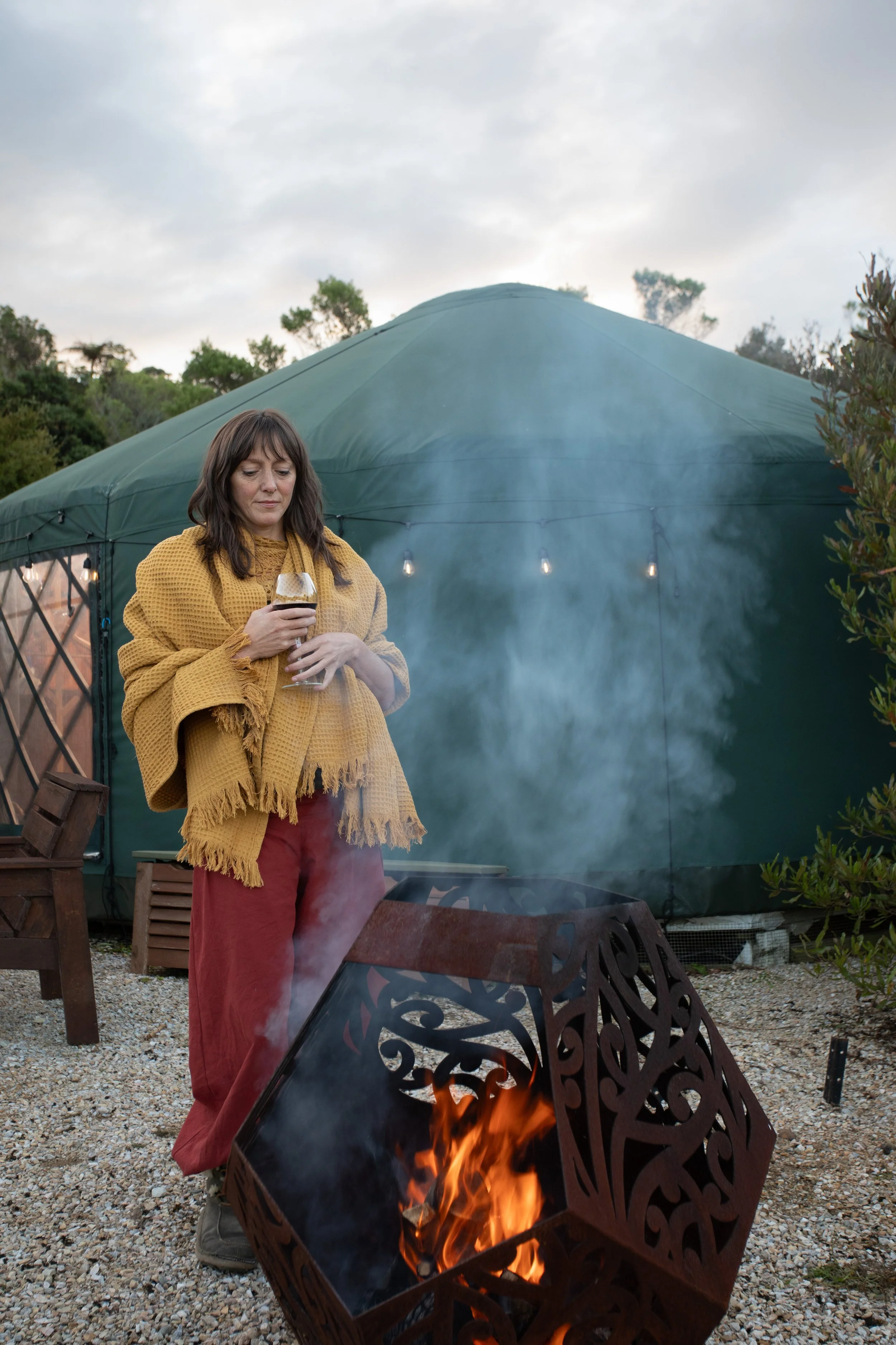 A woman standing outside near a fire pit with flames visible at the bottom of the image, holding a drink in a wine glass, with a large green tent and trees in the background during cloudy weather.
