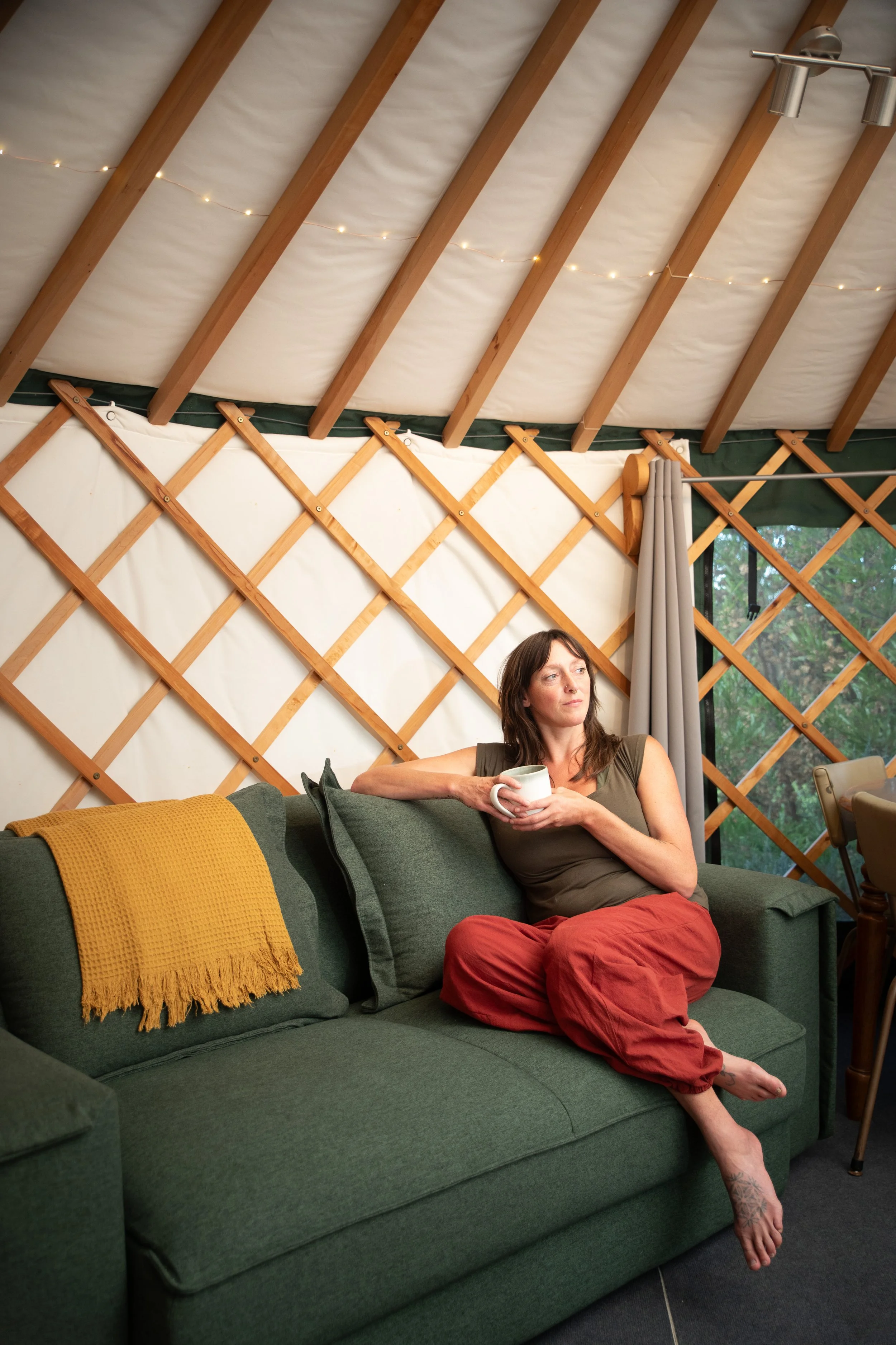 A woman sitting on a green couch in a cozy yurt and a window showing trees outside, holding a mug, wearing a gray shirt and red pants, with a yellow textured throw blanket on the couch.