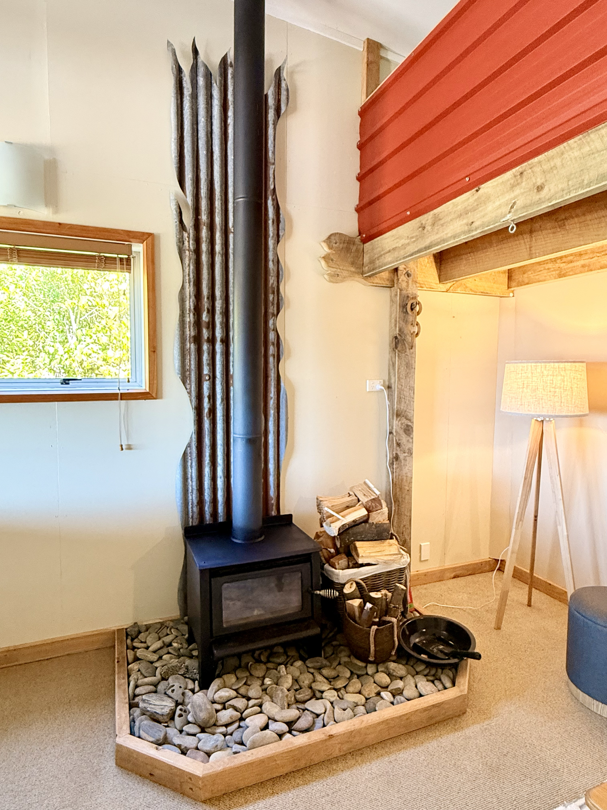 A cozy indoor space with a wood stove on a pebble base, surrounded by firewood and tools, next to a window and a loft with a red exterior wall.