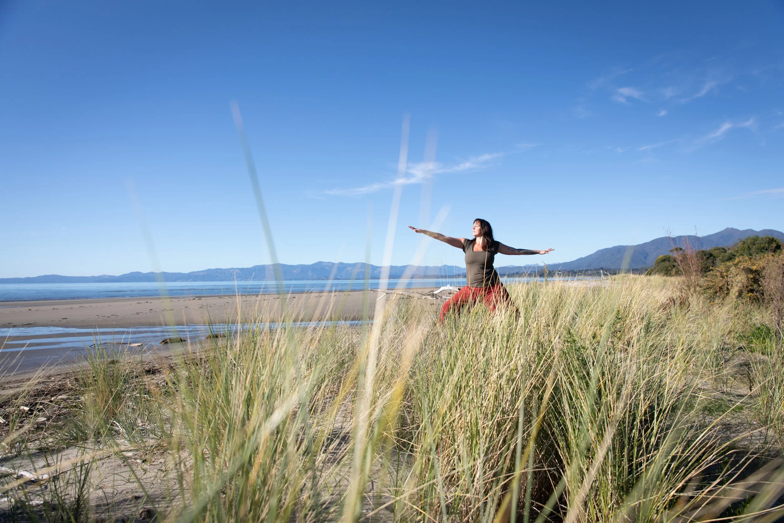 A woman practicing yoga outdoors on a grassy beach with mountains in the background, wearing a green shirt and red pants, with her arms extended outward and eyes closed under a clear blue sky.