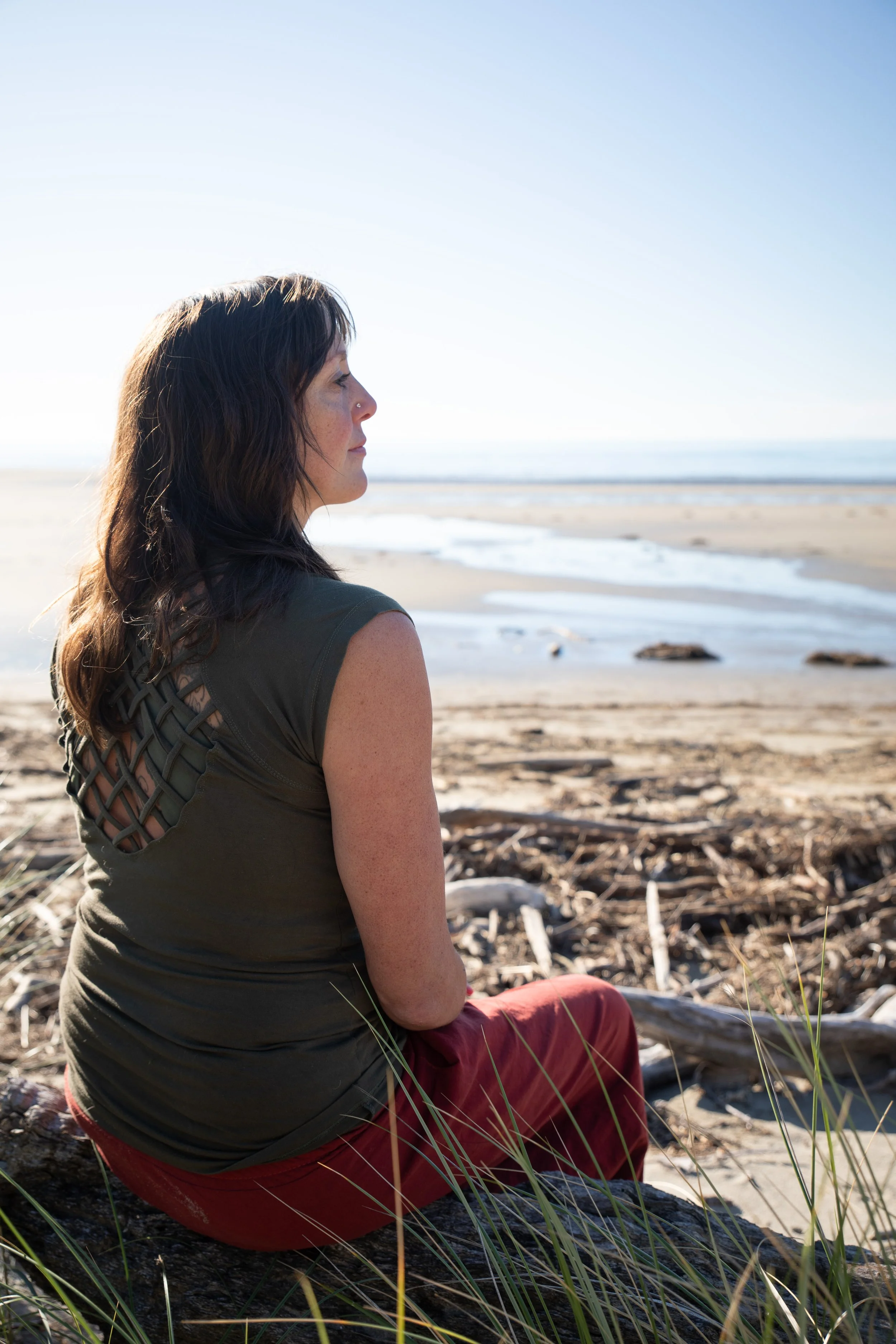 A woman with dark hair and a nose piercing sitting on a beach, facing the ocean with the sky above.