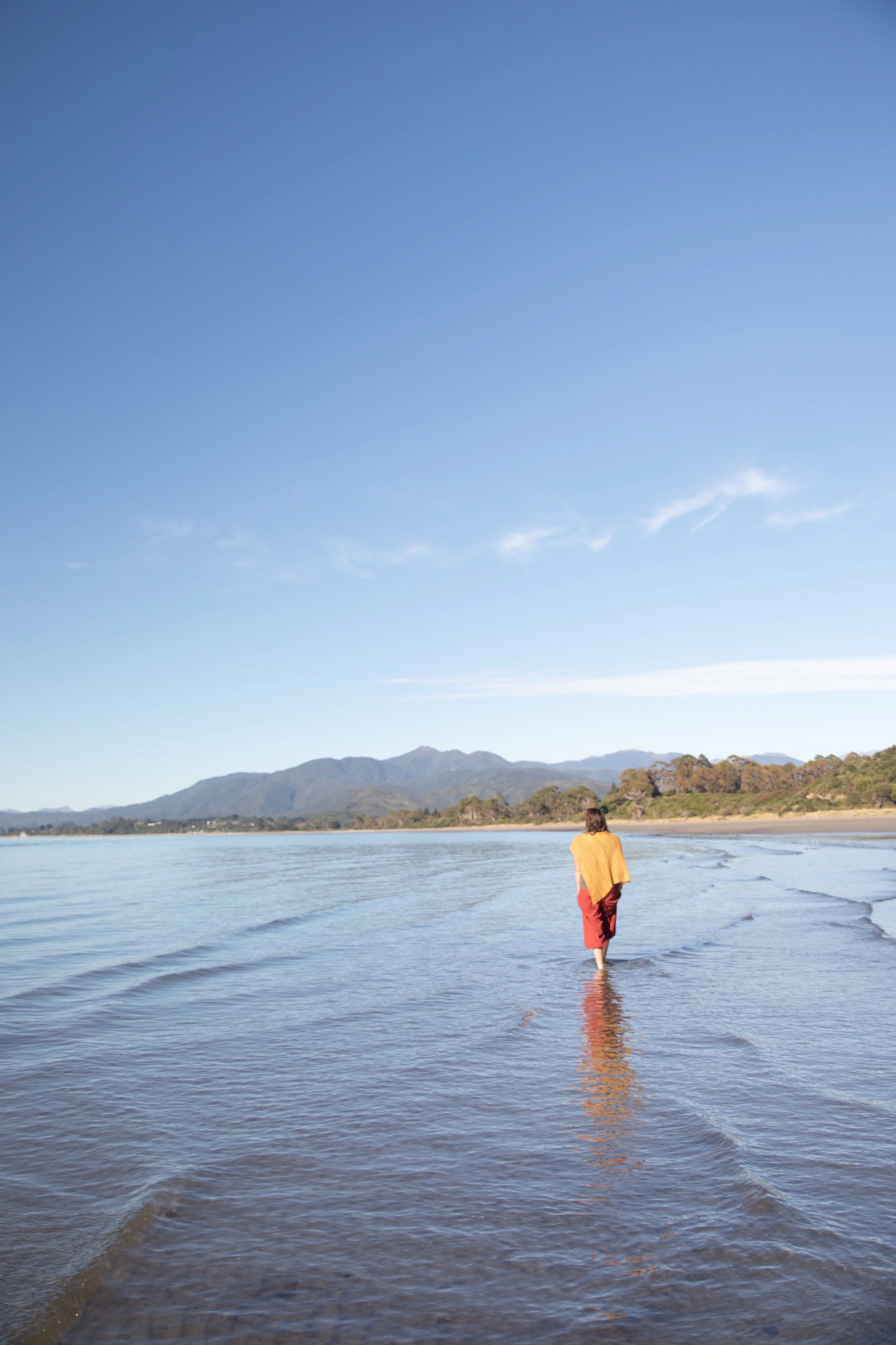 A person wearing red pants and a yellow top walking along a calm beach shoreline with mountains in the distance under a blue sky.