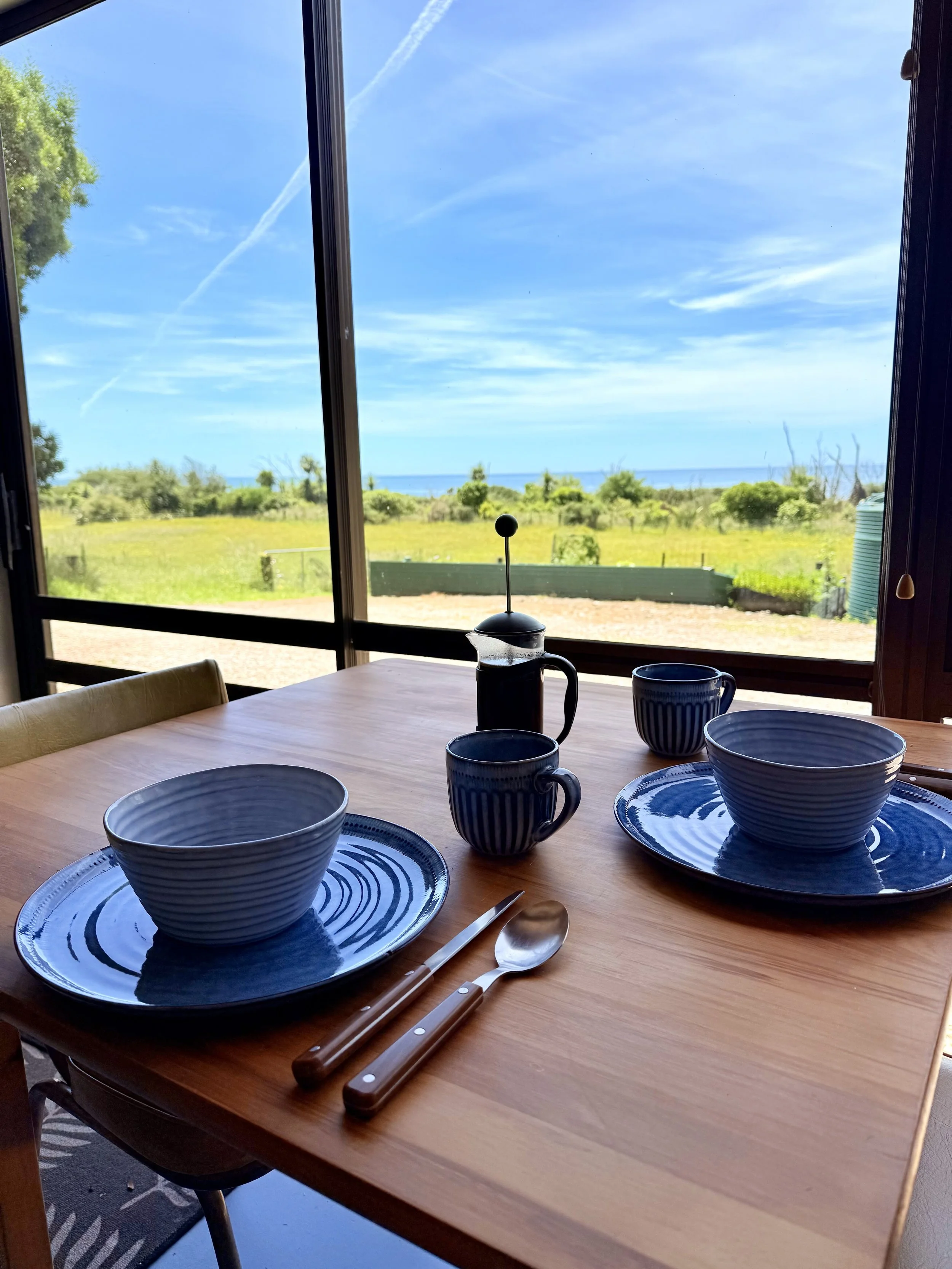 A wooden dining table set with two blue and white striped bowls on matching blue plates, a knife and spoon, and a French press with coffee, in front of a large window overlooking a scenic landscape with green fields, trees, and a distant view of the 