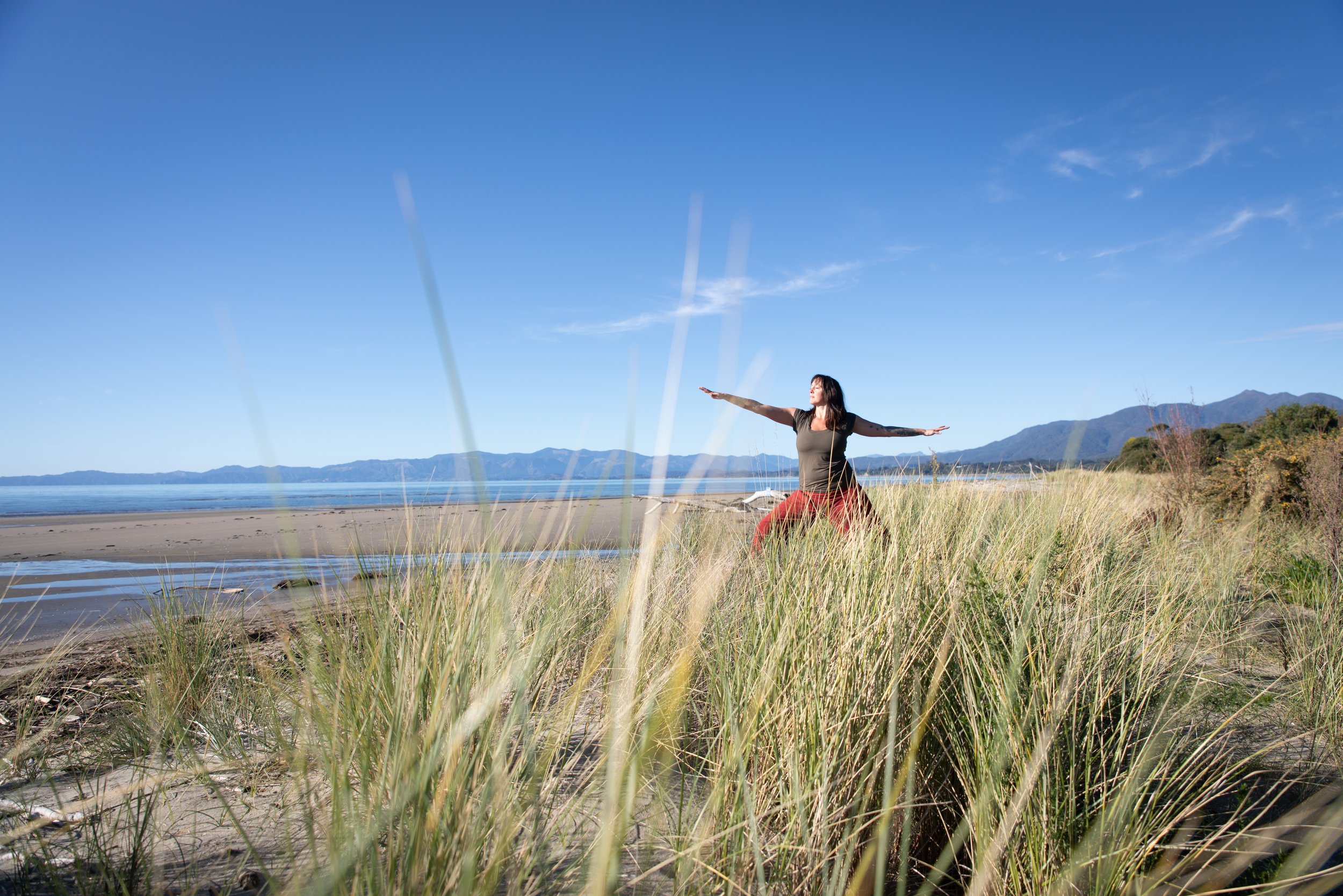 A woman practicing yoga outdoors on a grassy area near the beach, with mountains in the distance and a clear blue sky.