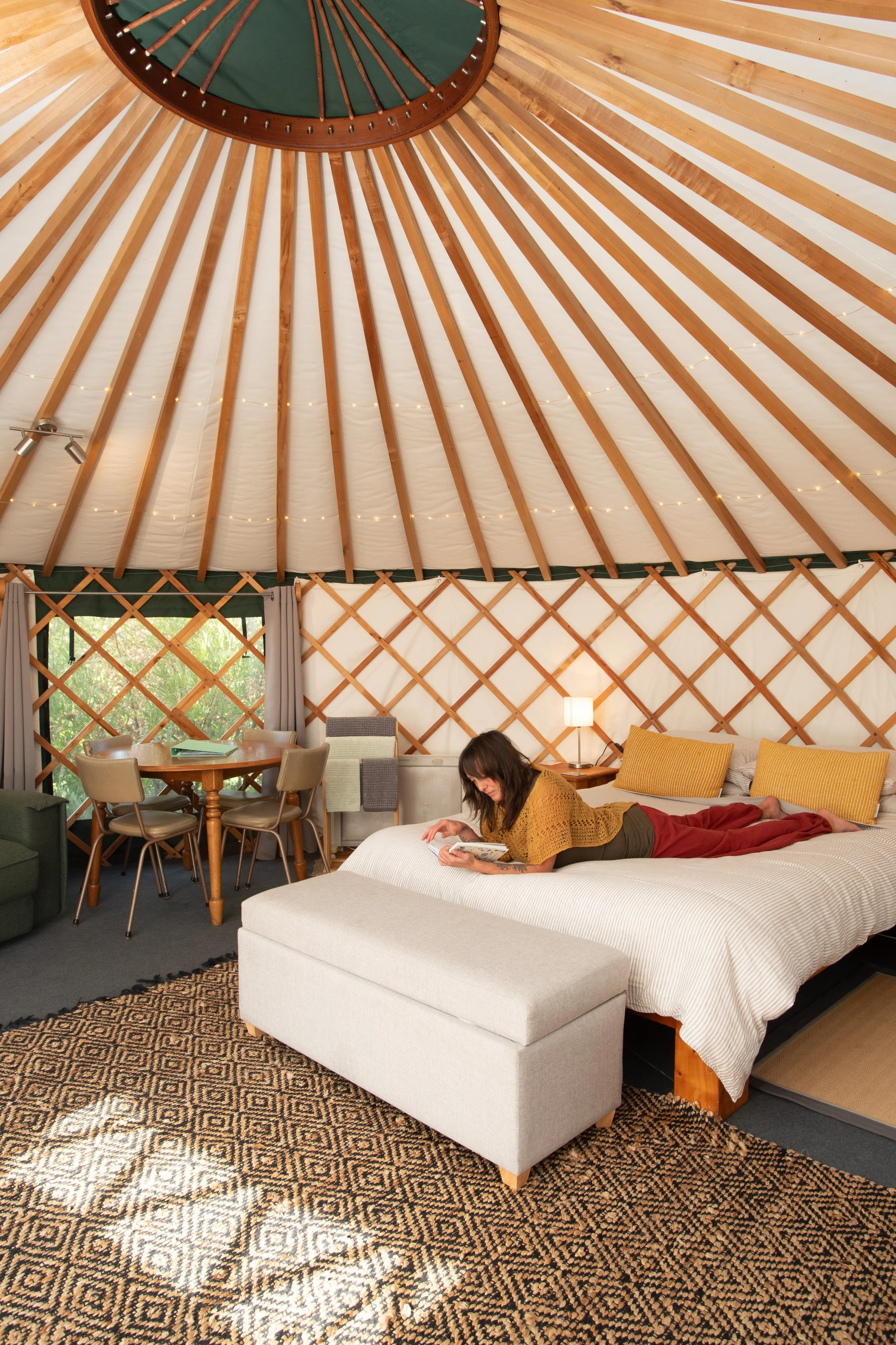 Interior of a cozy yurt with a woman lying on a bed reading a book, wooden beams on the ceiling, and a sitting area with a dining table and chairs.