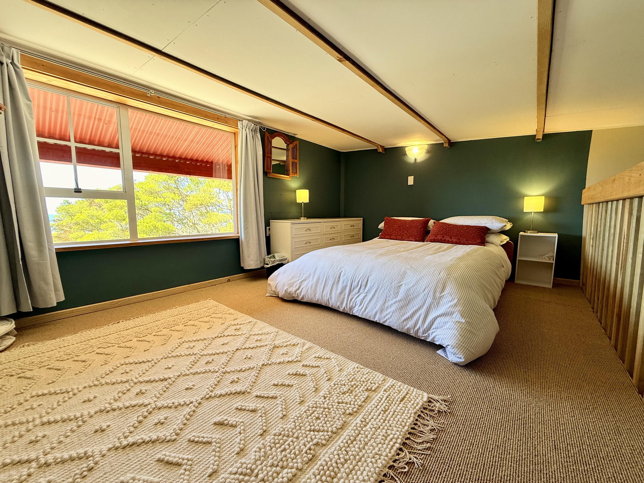 A bedroom with a large window showing trees outside, a bed with white bedding and red pillows, a white nightstand with a yellow lamp, a white dresser, and a wall-mounted mirror.