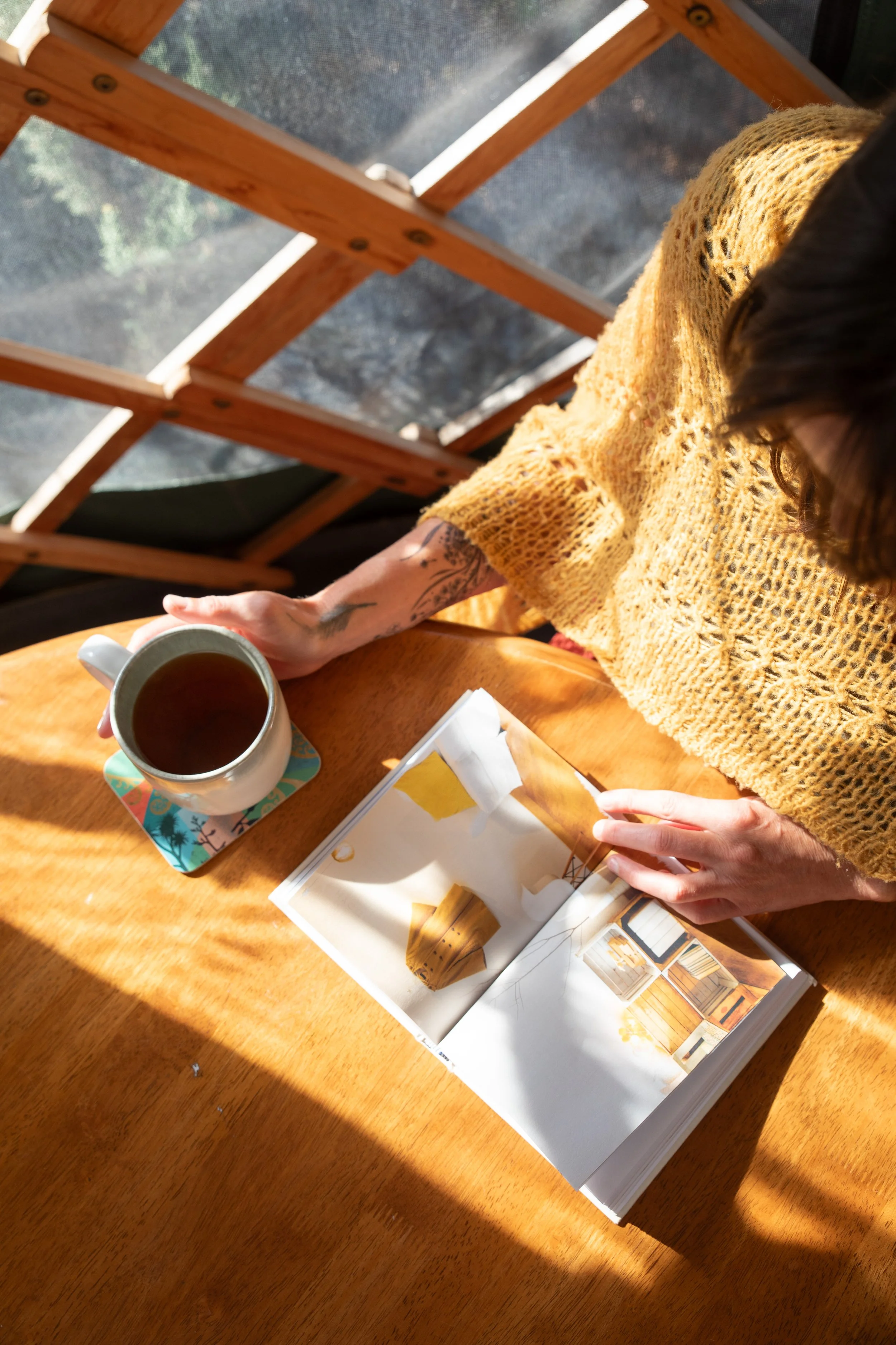 A person wearing a yellow knitted sweater is sitting at a wooden table, holding a coffee mug. There is an open interior design magazine with pictures of rooms on the table. The person has a tattoo on their left arm and is in a space with sunlight streaming in, with a wooden framed structure and a screen in the background.