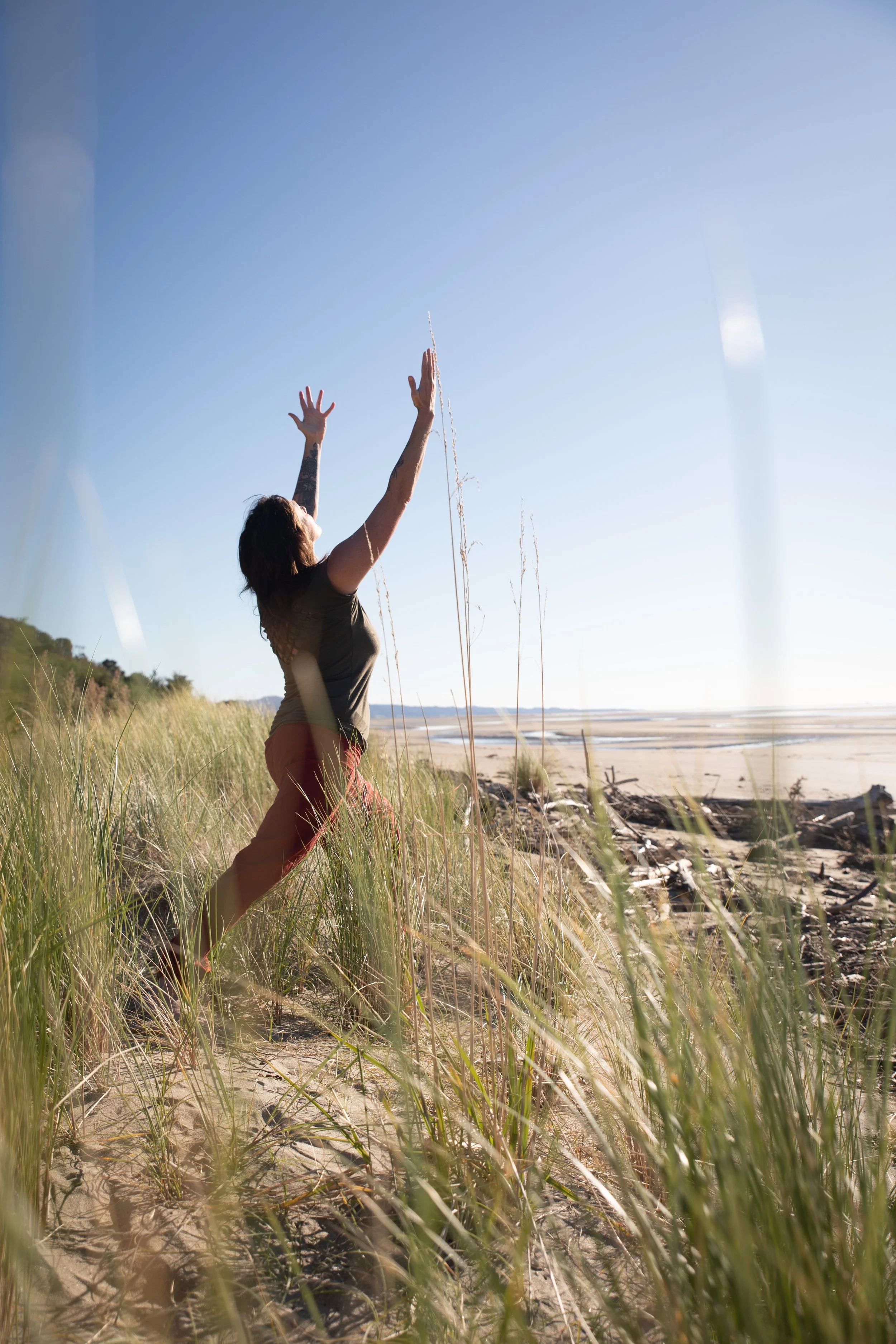 A woman with tattoos on her arms and dark hair, standing in tall green grass on a sandy beach, reaching up in a yoga pose towards a clear blue sky with her arms extended.