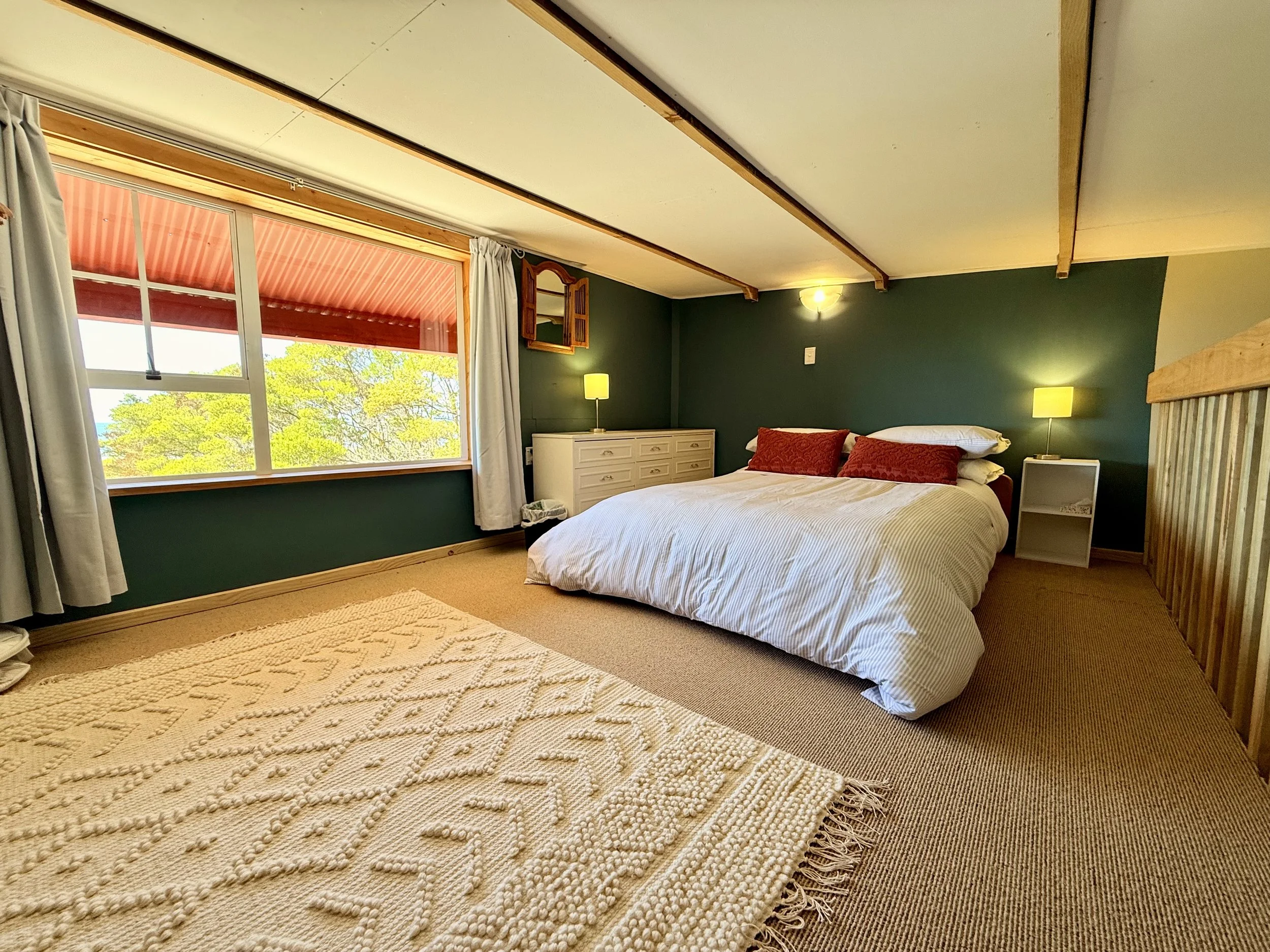 A cozy bedroom with a large window showing green trees outside, a white bed with red pillows, two green lamps on white nightstands, and light-colored curtains and carpet.