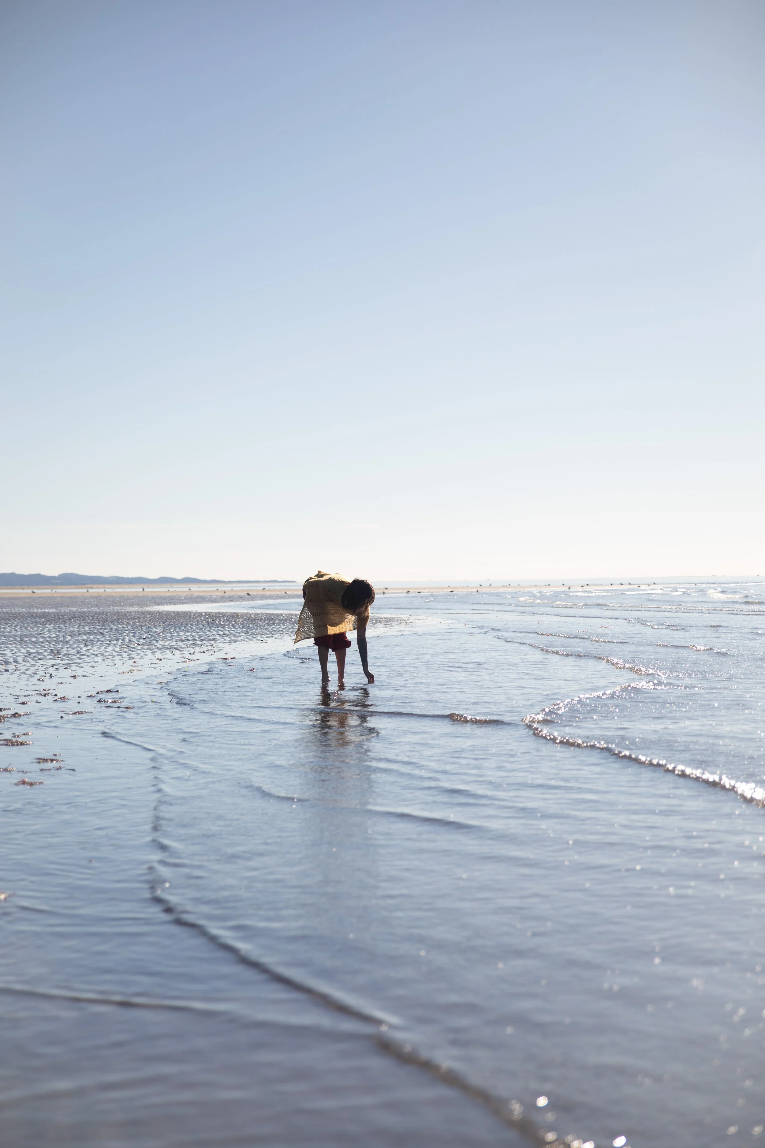 Person in a beige sweater and red pants bending down on a sandy beach near the water, with the ocean extending to the horizon under a clear sky.