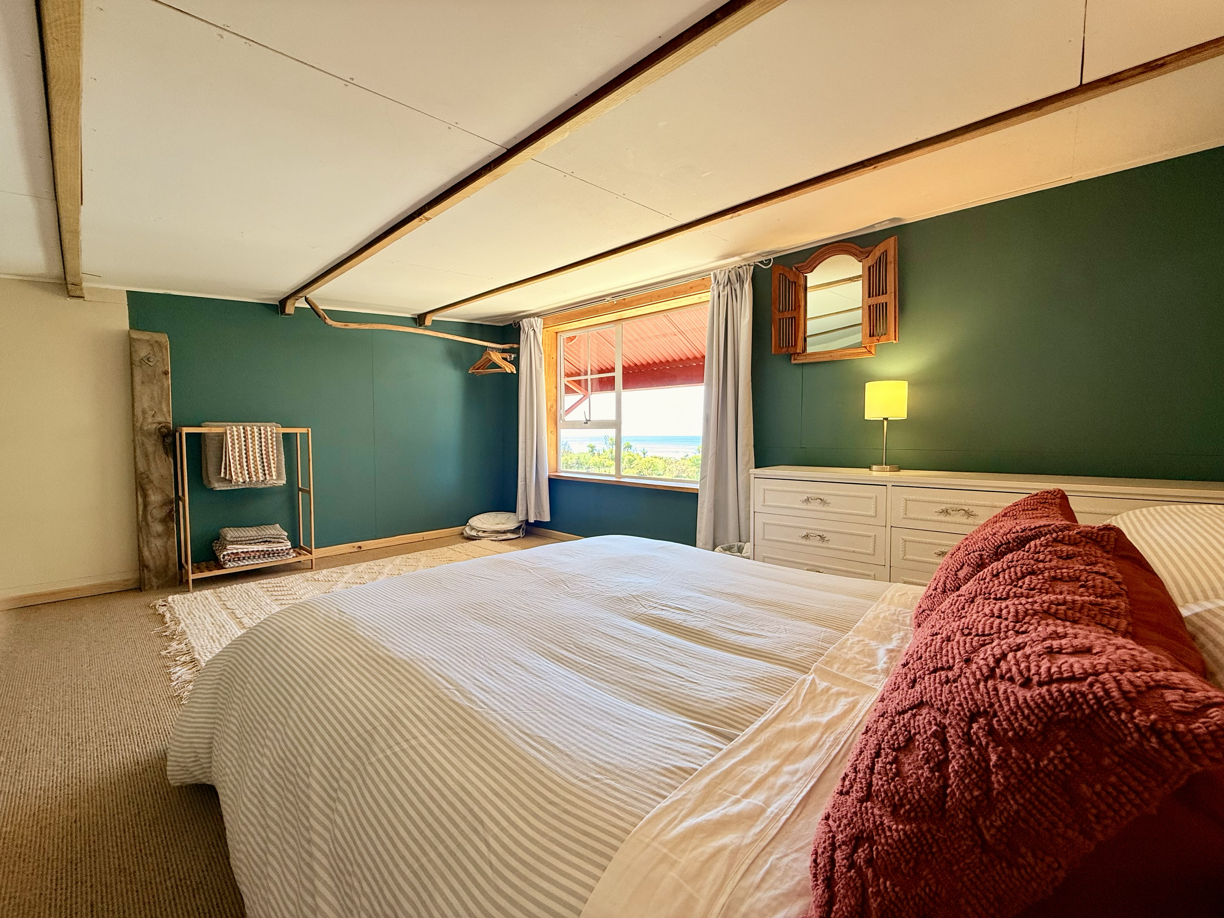 Cozy bedroom with a large bed, striped bedding, and red pillows, a white dresser with a lamp, a window with white curtains showing an outdoor sea view, wooden wall accents, and minimalist decor.