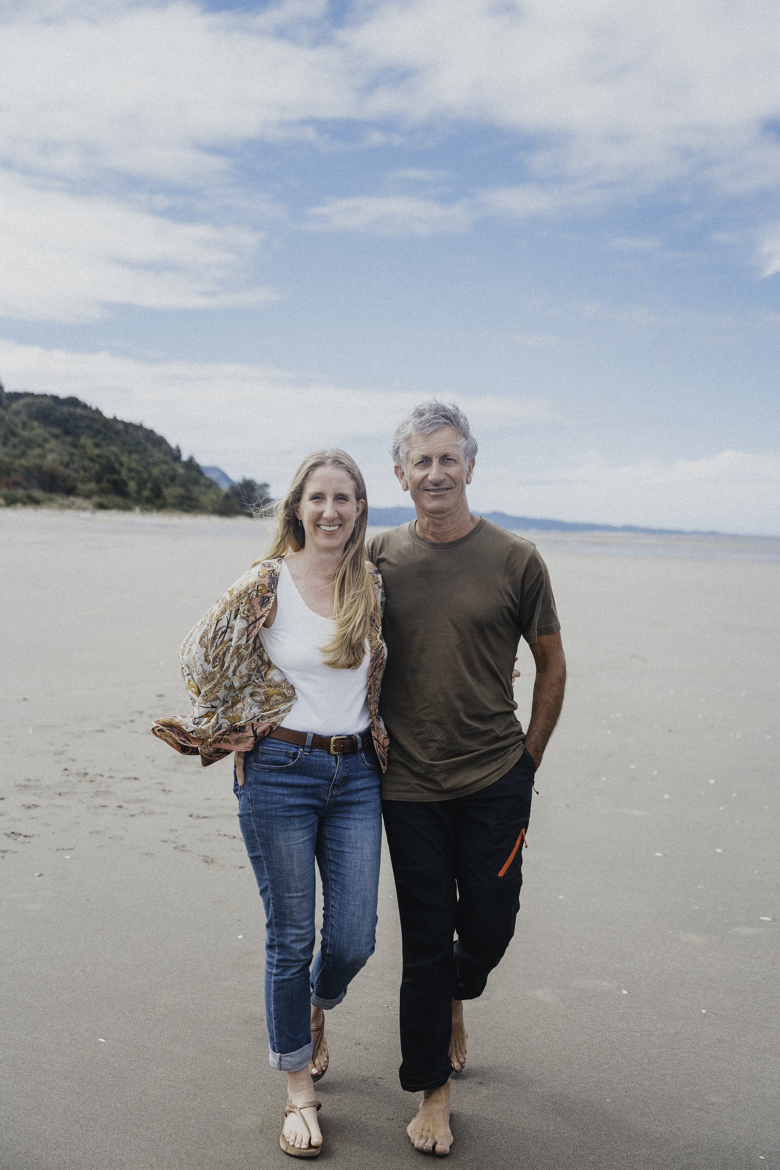 A smiling woman and man walking barefoot on a sandy beach with ocean and distant hills in the background.