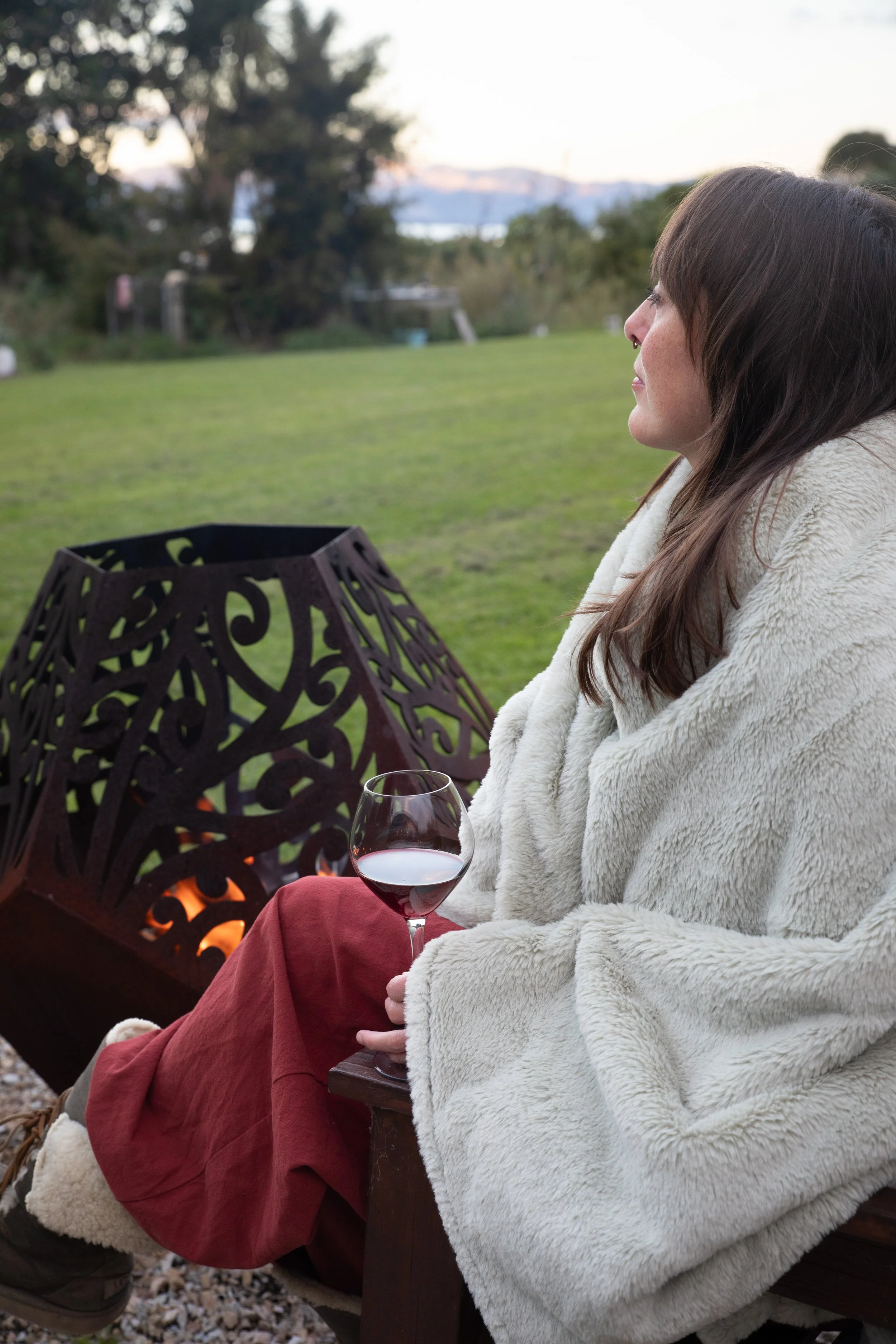 A woman sitting outdoors near a fire pit, holding a glass of red wine, wrapped in a soft blanket and red pants, with a lawn, trees and the ocean in the background.