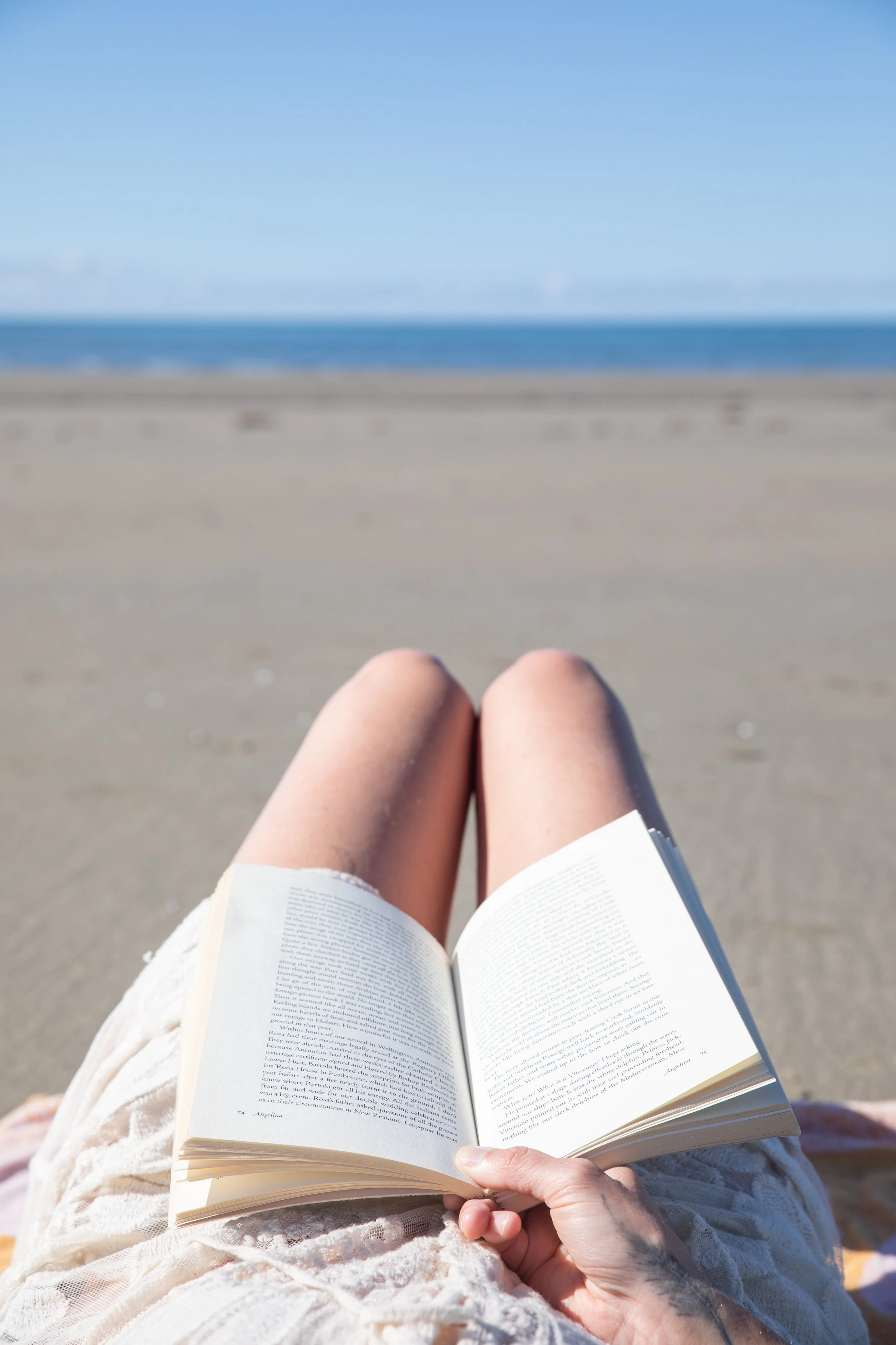 Person sitting on a beach reading a book with ocean and blue sky in the background.