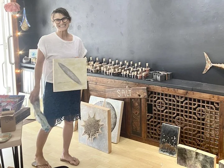 Woman smiling holding a painting in an art studio, surrounded by art pieces, decorative items, and a shelf labeled 'salt' with jars on it.