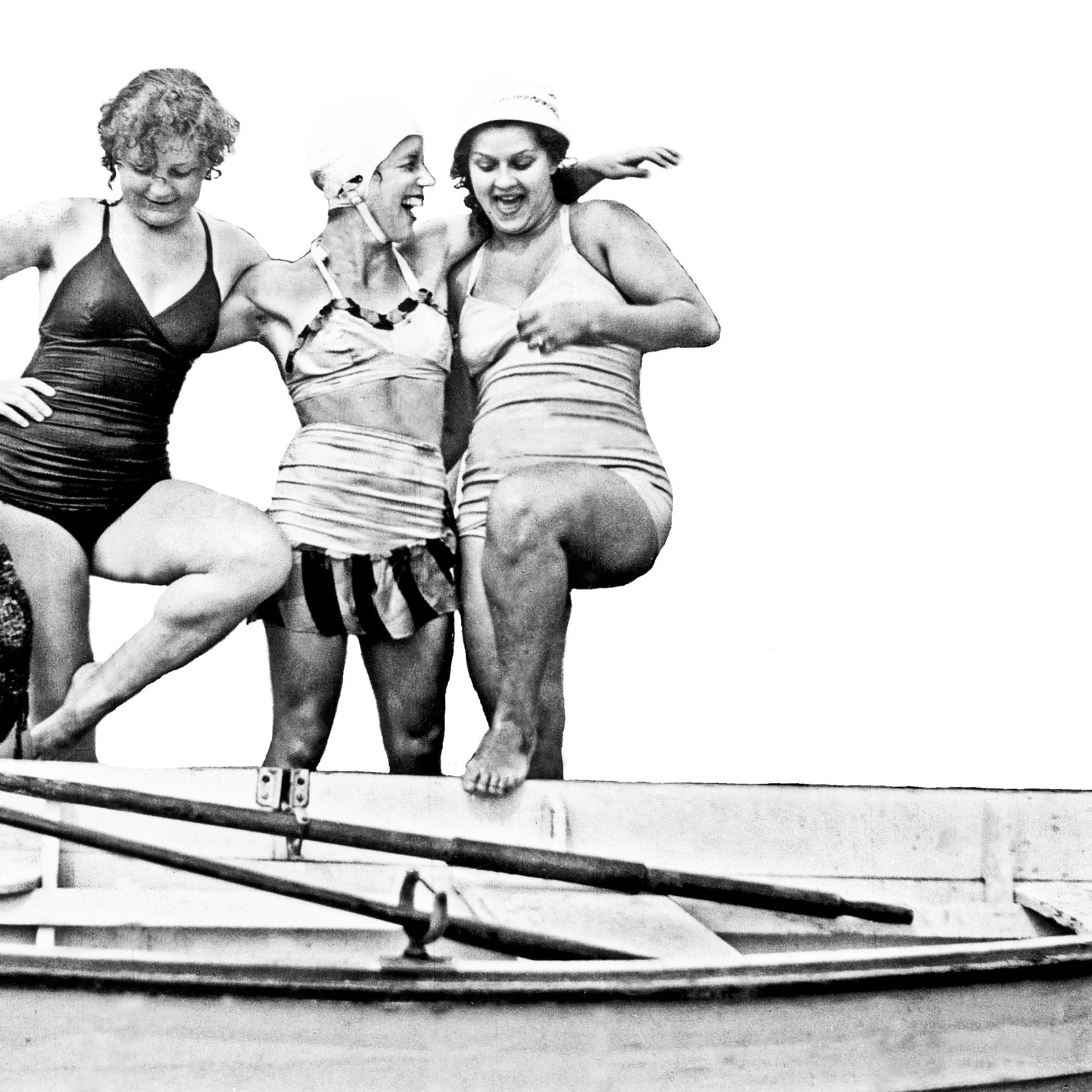 Three women in vintage swimsuits and swim caps pose together, playfully lifting their legs while standing on a boat.