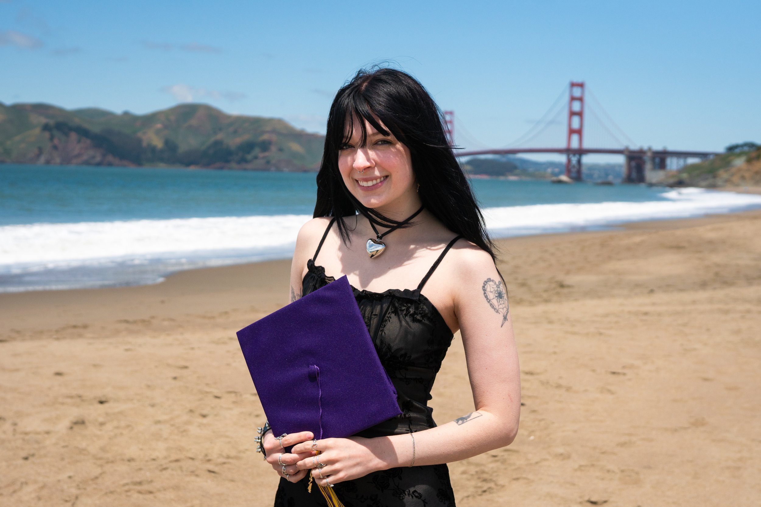 A young woman with dark hair in a black dress holding a diploma on a beach with a bridge and hills in the background.