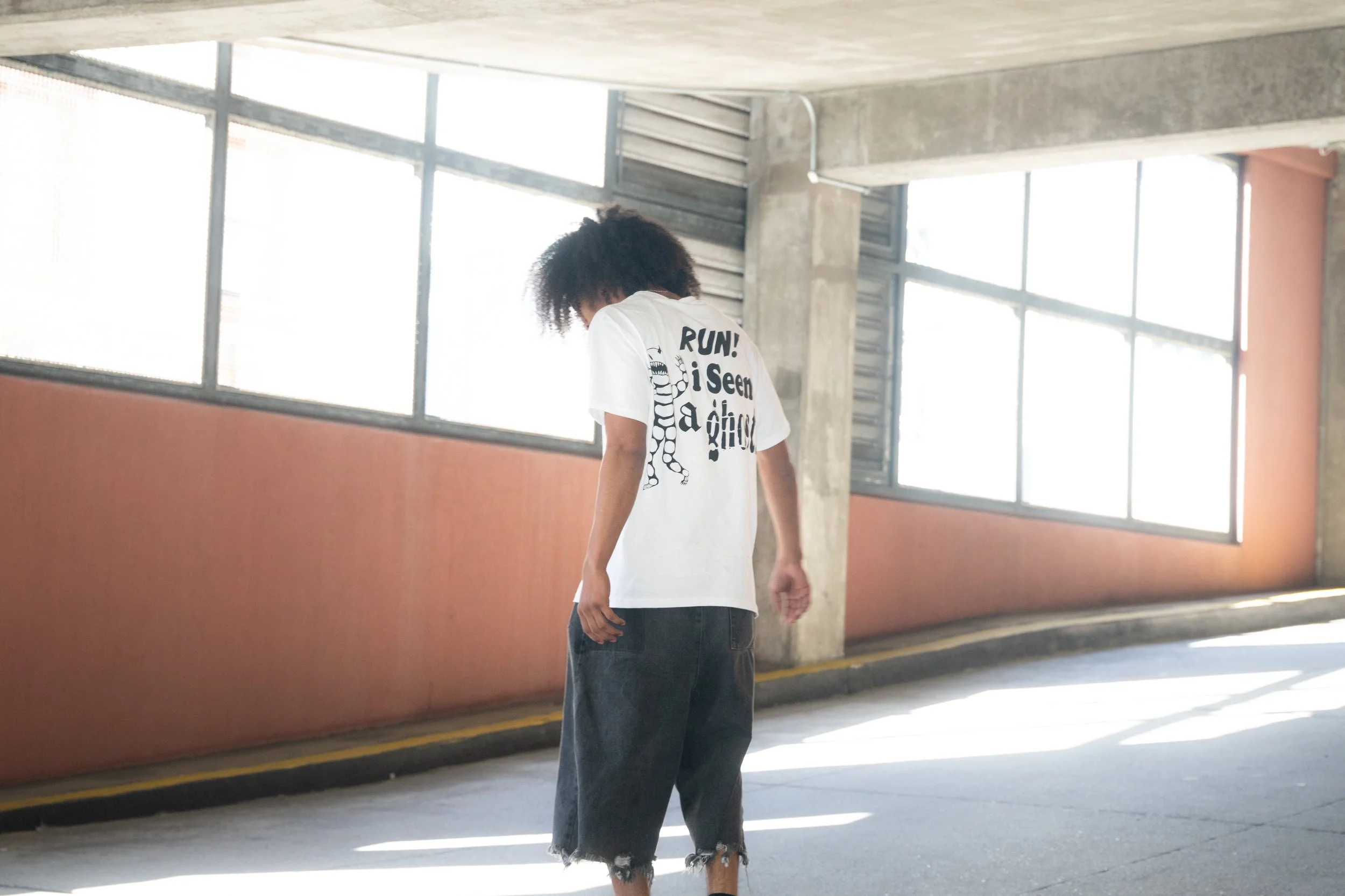Person with curly hair walking in a parking garage, wearing a white T-shirt with text and black ripped jeans.