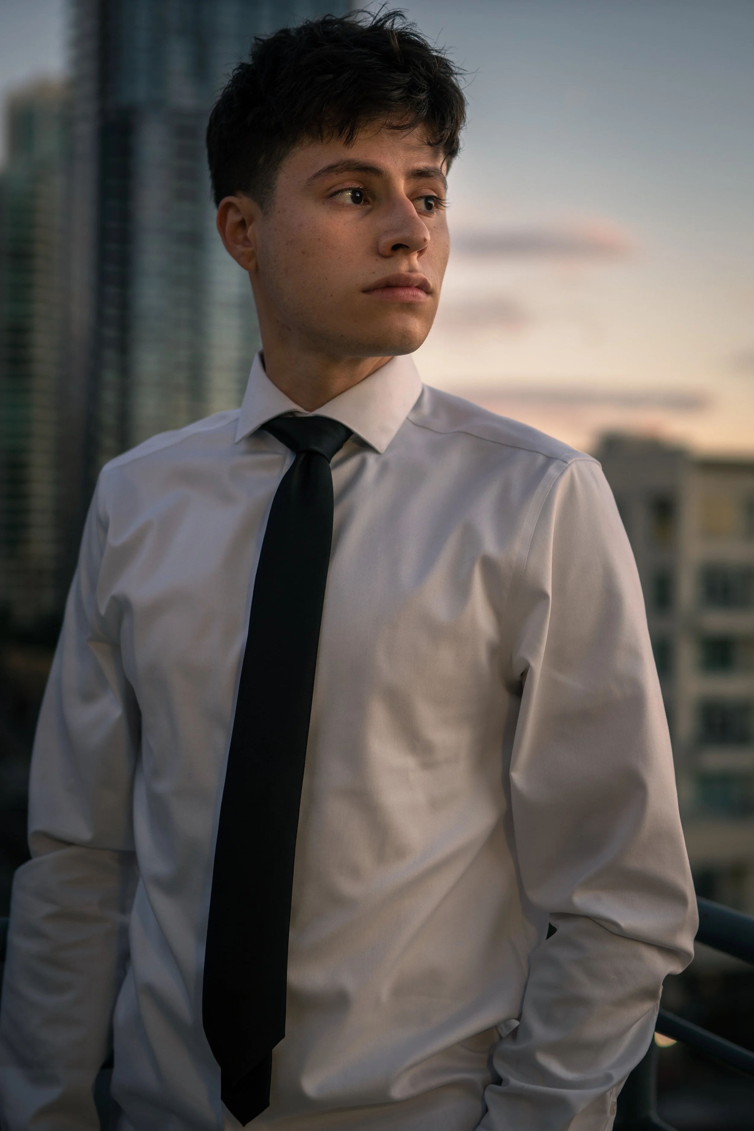 Young man in a white dress shirt and black tie standing on a balcony in an urban area during sunset.