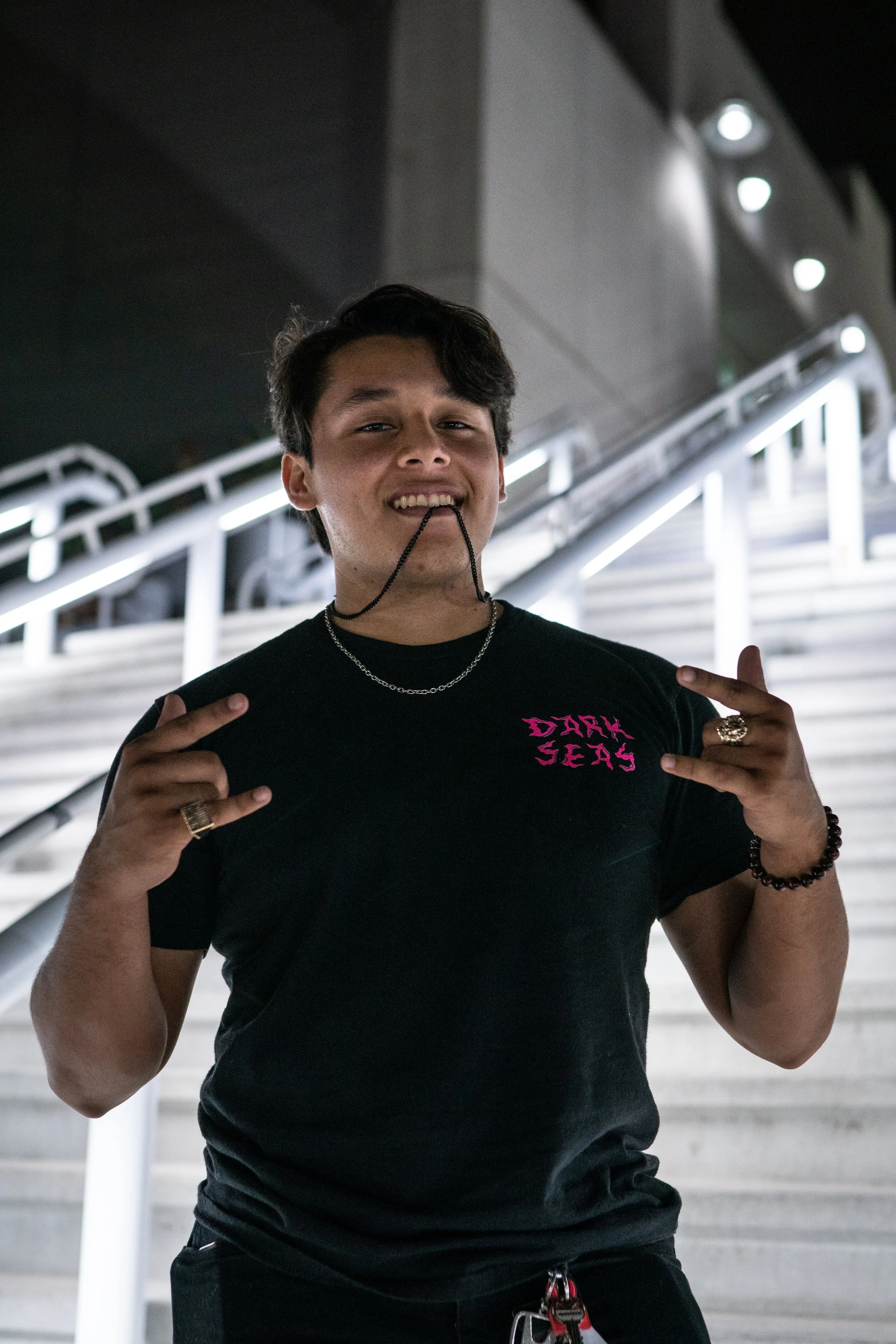 A young man smiling at the camera, wearing a black t-shirt with pink text that says 'DARK SEAS,' making rock hand gestures, standing in front of a staircase at night with bright lights.