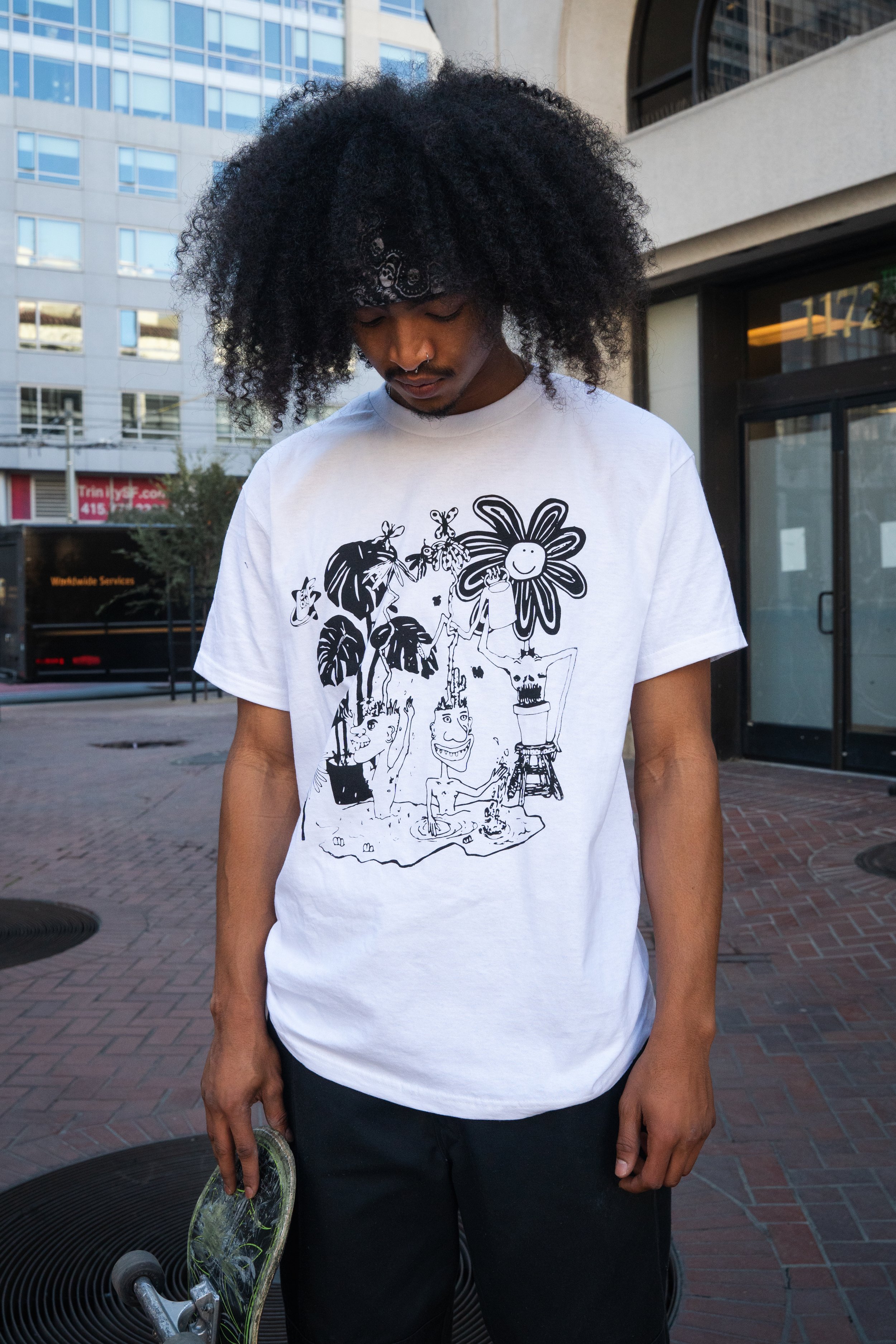 A young man with curly black hair and a bandana, wearing a white graphic t-shirt and black pants, standing on a city sidewalk holding a skateboard, with modern office buildings in the background.