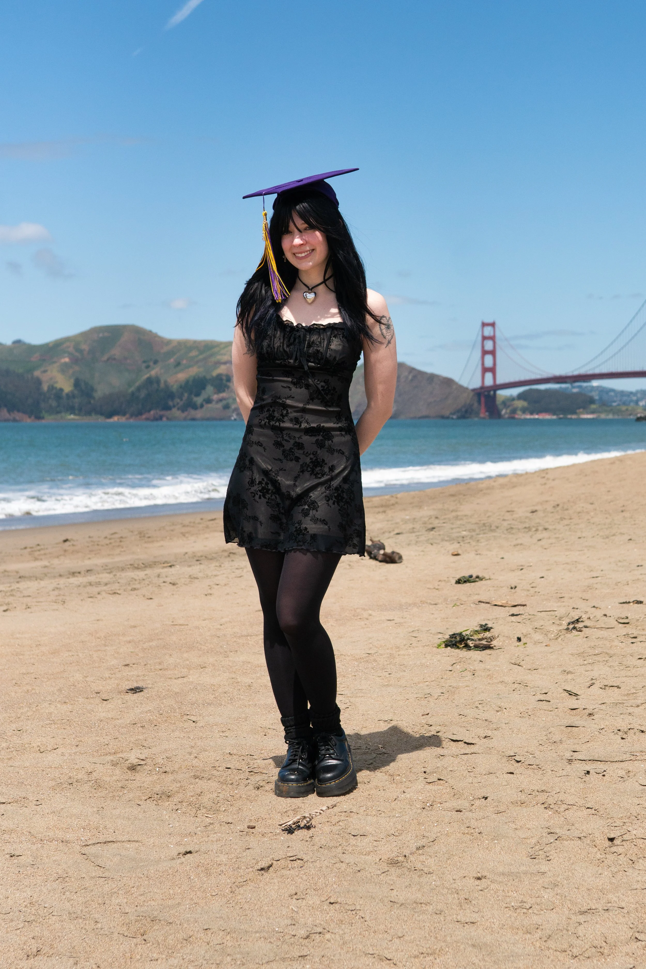 A young woman with black hair wearing a black dress, black tights, and black boots, smiling and standing on a sandy beach with the Golden Gate Bridge in the background.