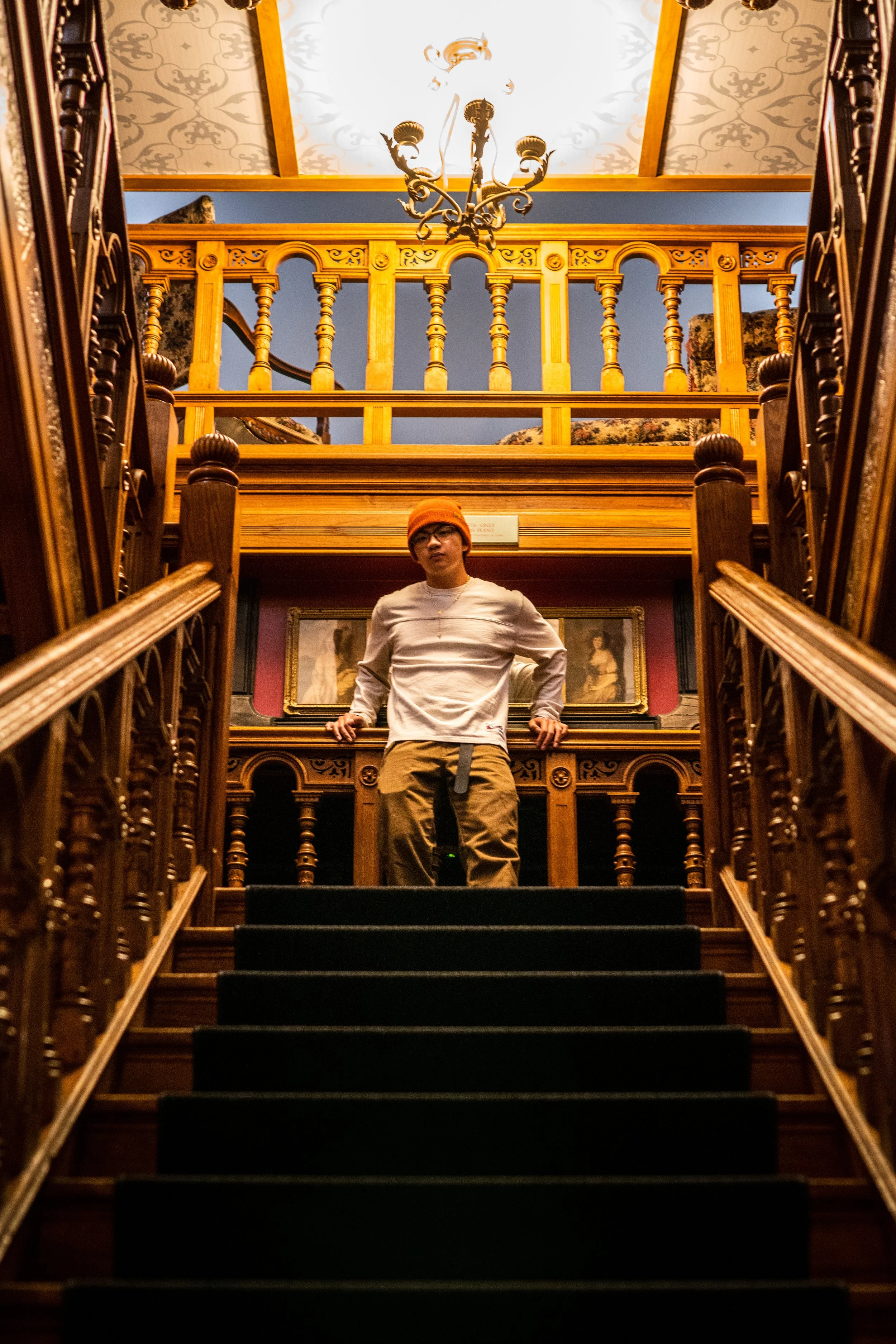 A person standing on a staircase in a room with ornate wooden railings and framed paintings, looking down at the camera, wearing an orange beanie, glasses, a white long sleeve shirt, and khaki pants.