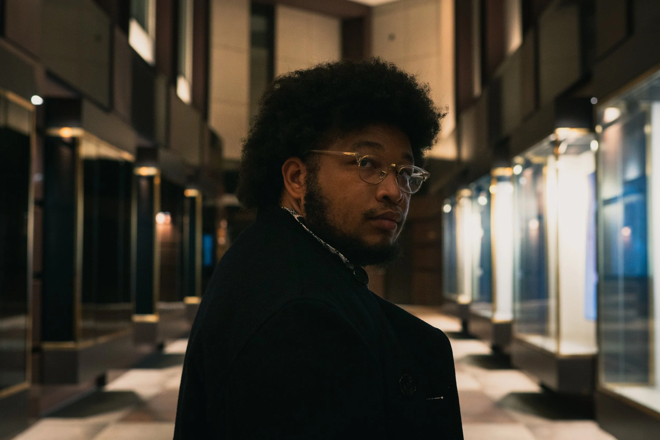 A man with glasses and headphones around his neck standing in a dimly lit indoor space, possibly a hallway or lobby, with illuminated display cases along the sides.