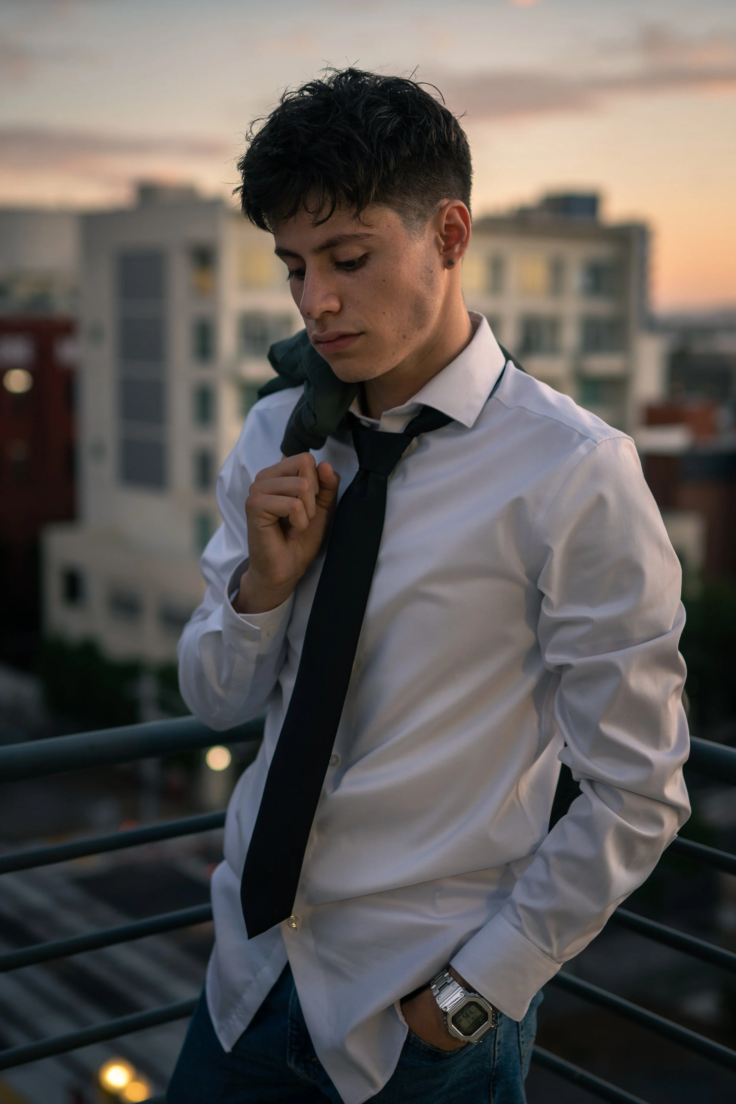 Young man in a white shirt, black tie, and jeans standing on a balcony at dusk, holding a jacket over his shoulder and looking down.