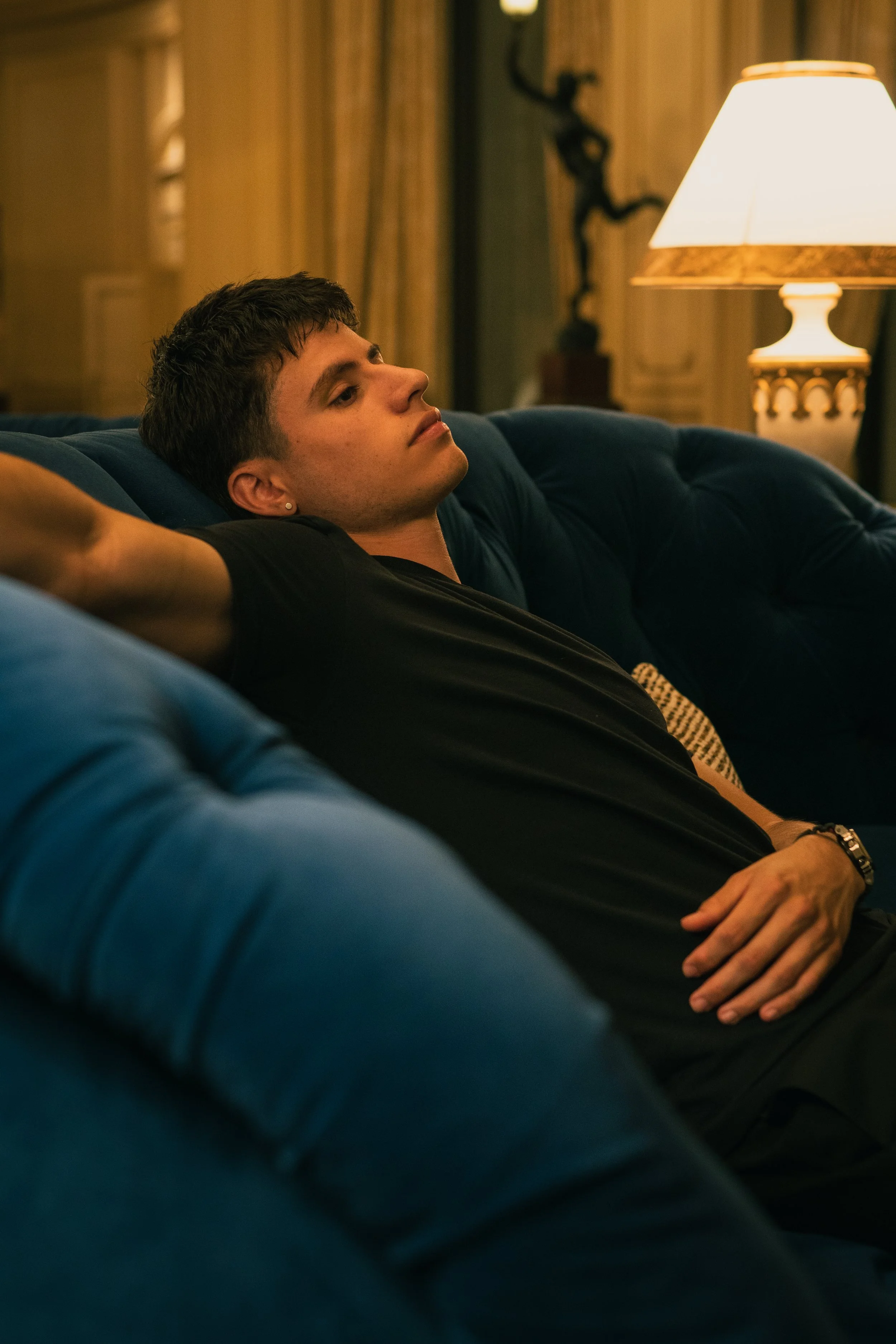Young man lying on a blue sofa with his arm outstretched, relaxing in a warmly lit room near a lamp and curtains.